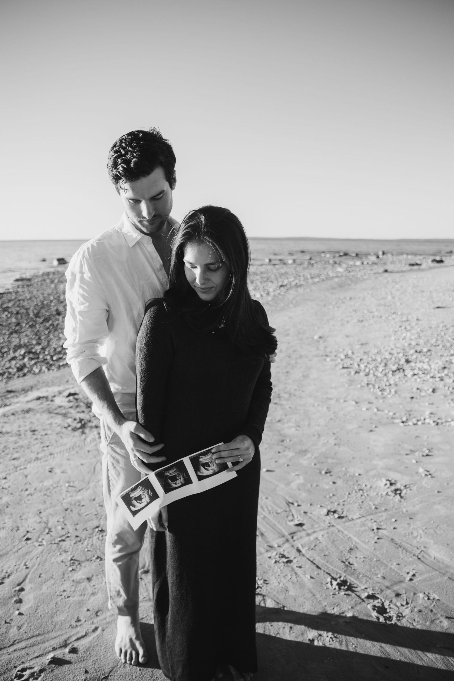 Black and white photo of an expecting couple embracing on a beach while holding ultrasound images, a maternity announcement.