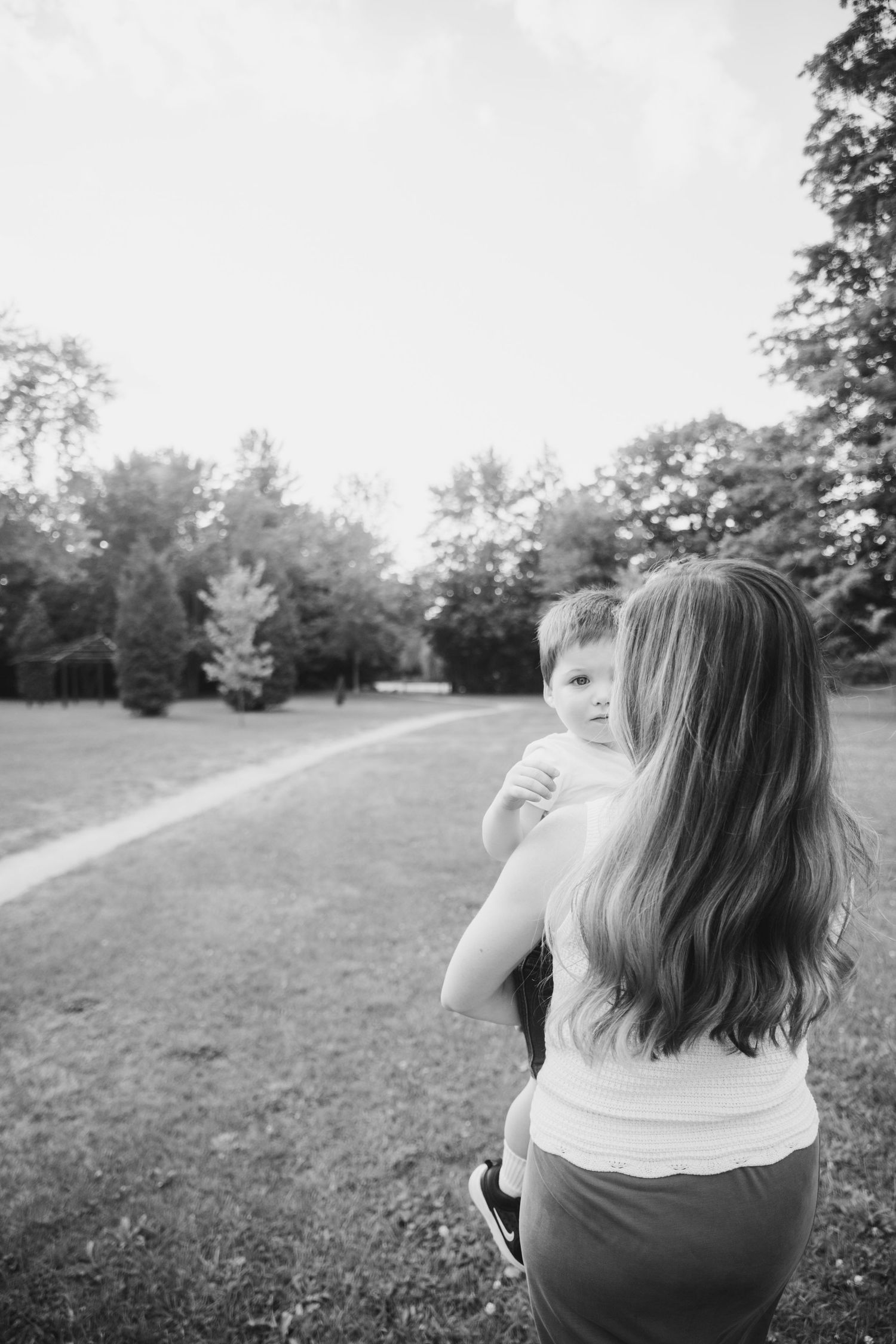 Black and white photo of a woman with long hair carrying a young child across a grassy park path lined with trees.