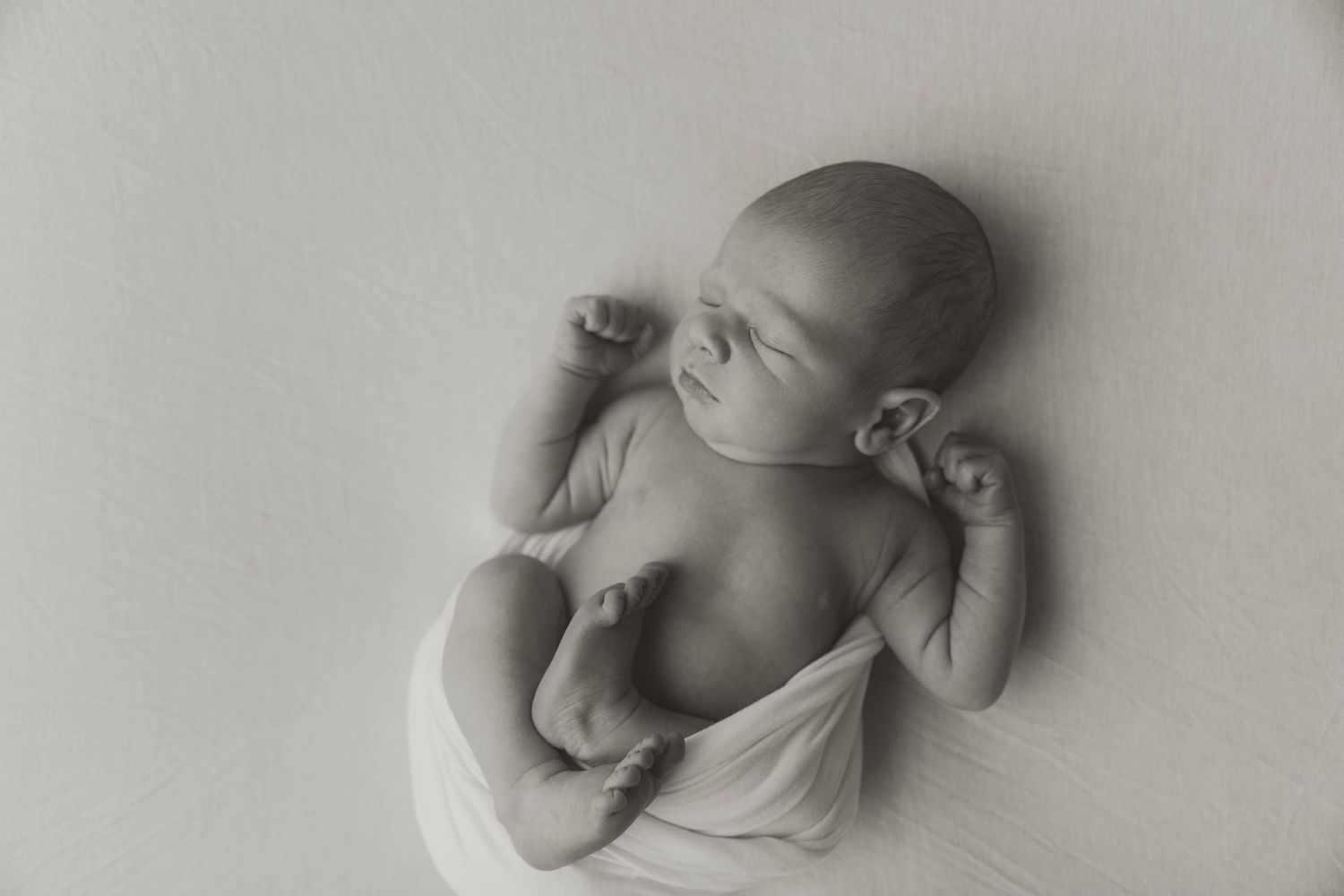 Black and white photograph of a sleeping baby swaddled in white fabric lying on their back.