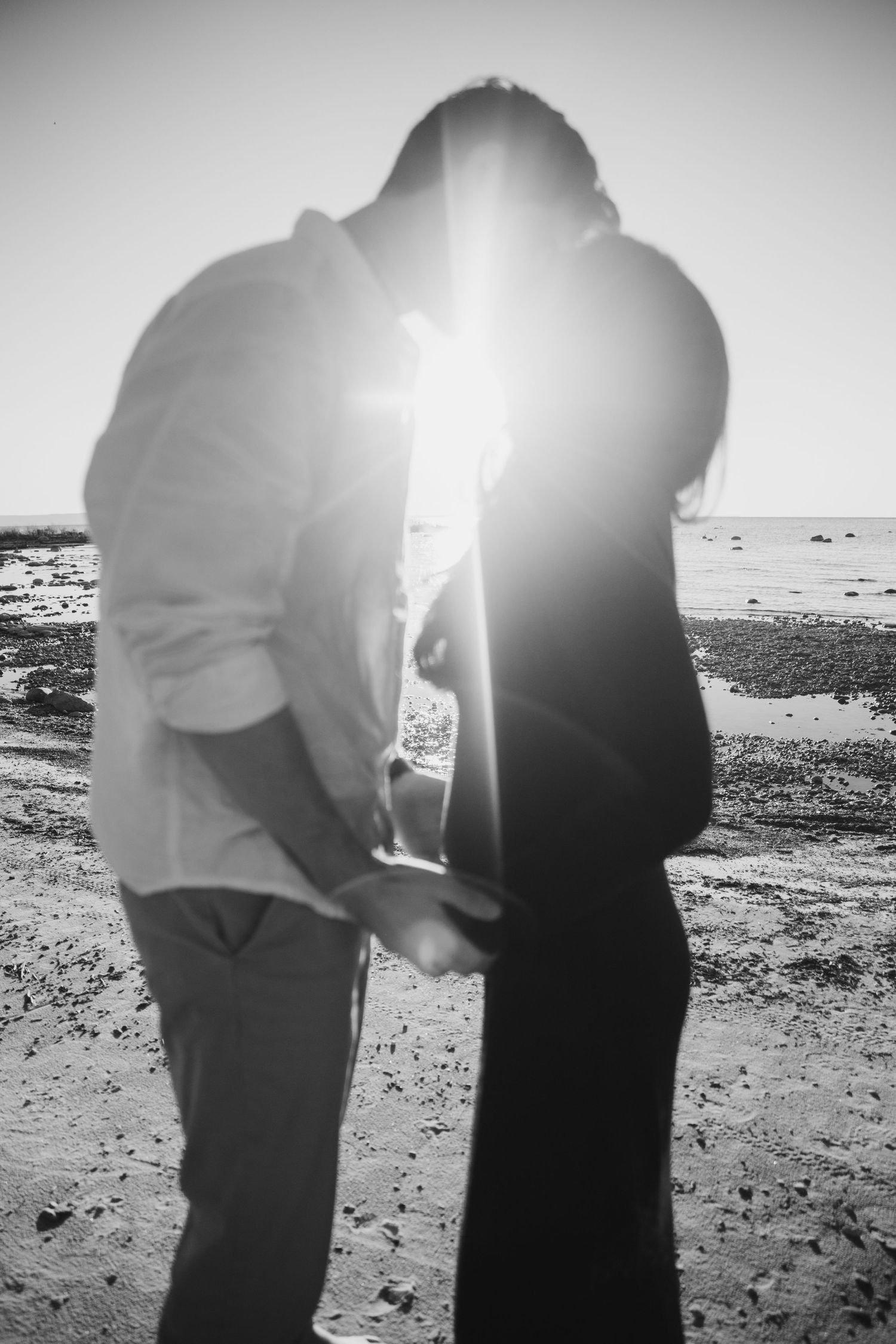 Silhouetted couple embracing on a rocky beach in black and white, with bright sunlight flaring between them at the shoreline.