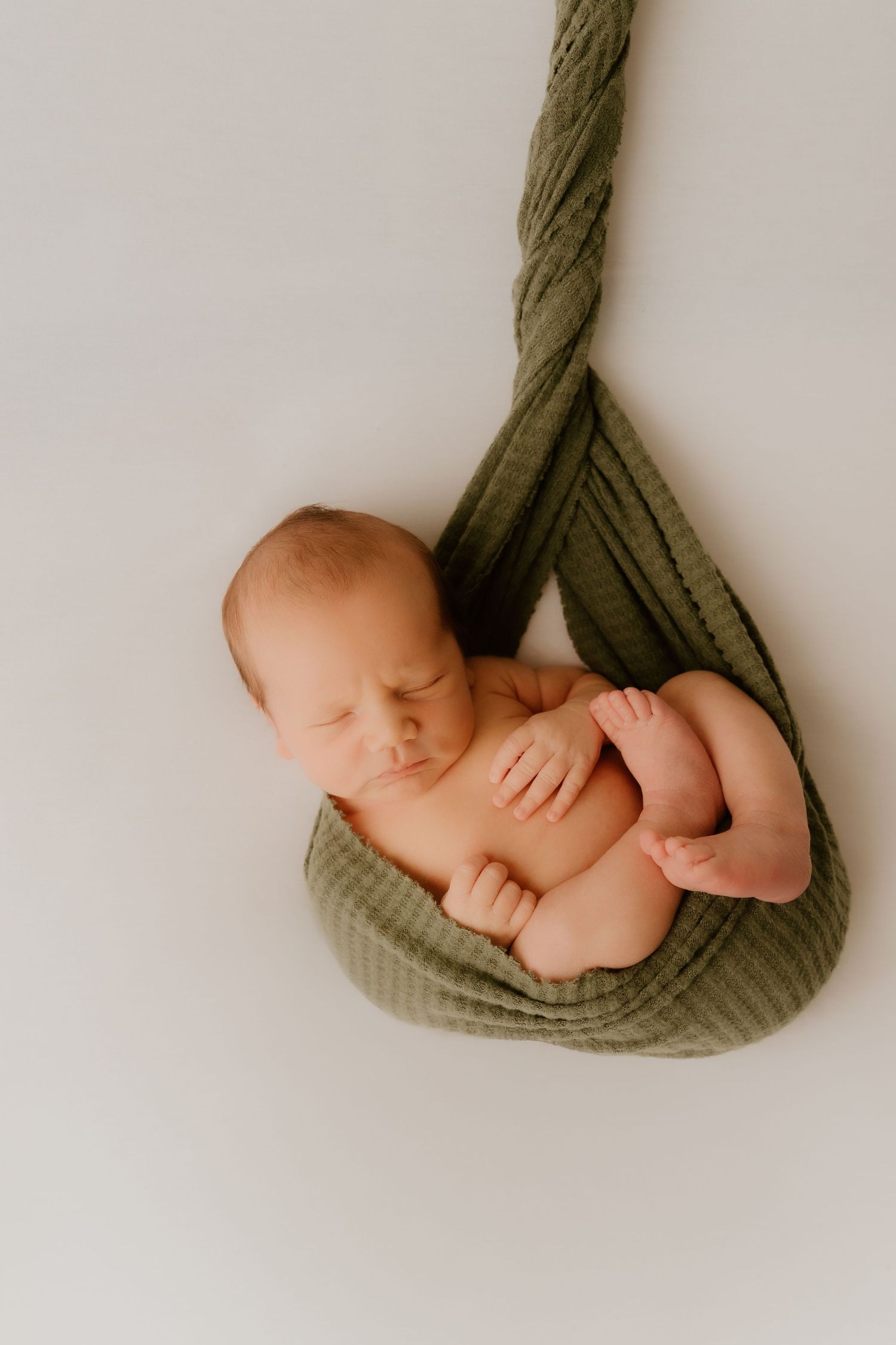 Newborn baby sleeping peacefully in a green knit wrap hammock against a white background.
