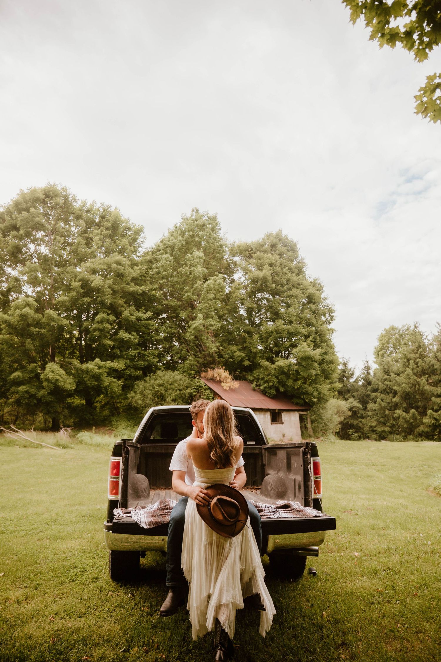 A couple sits together on the tailgate of a pickup truck in a sunny meadow.