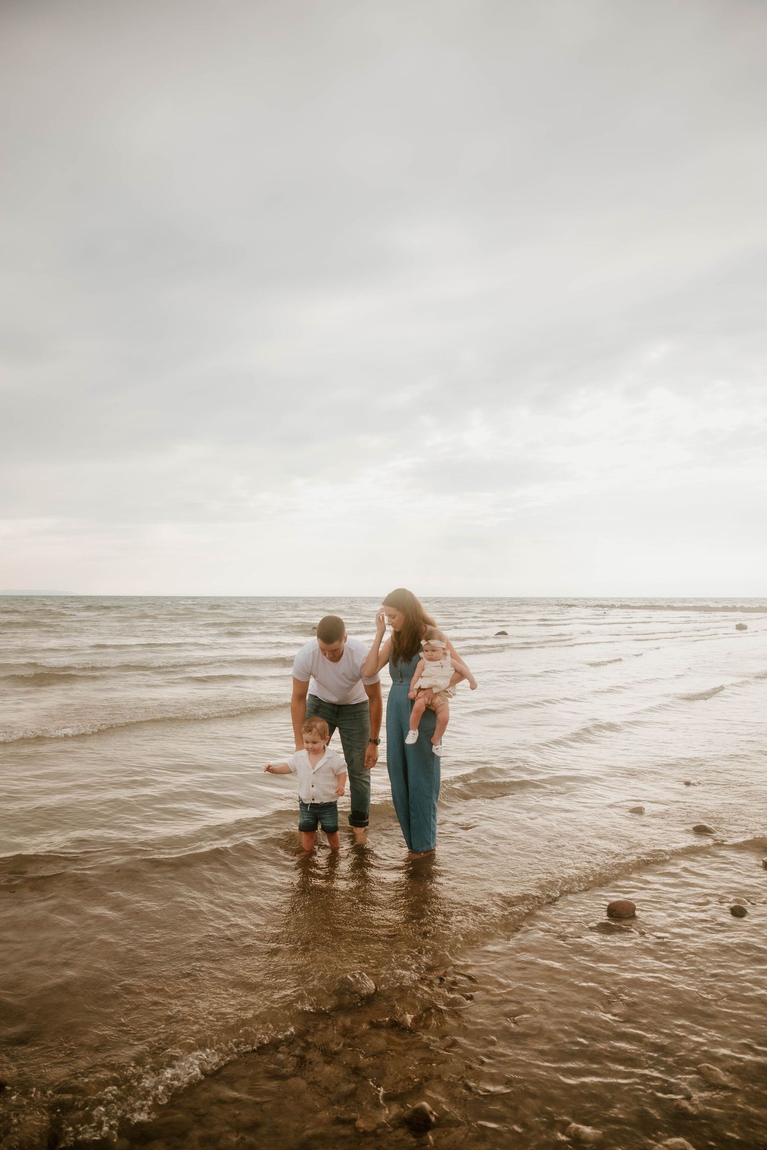 Family wading together in shallow ocean waves on an overcast day.