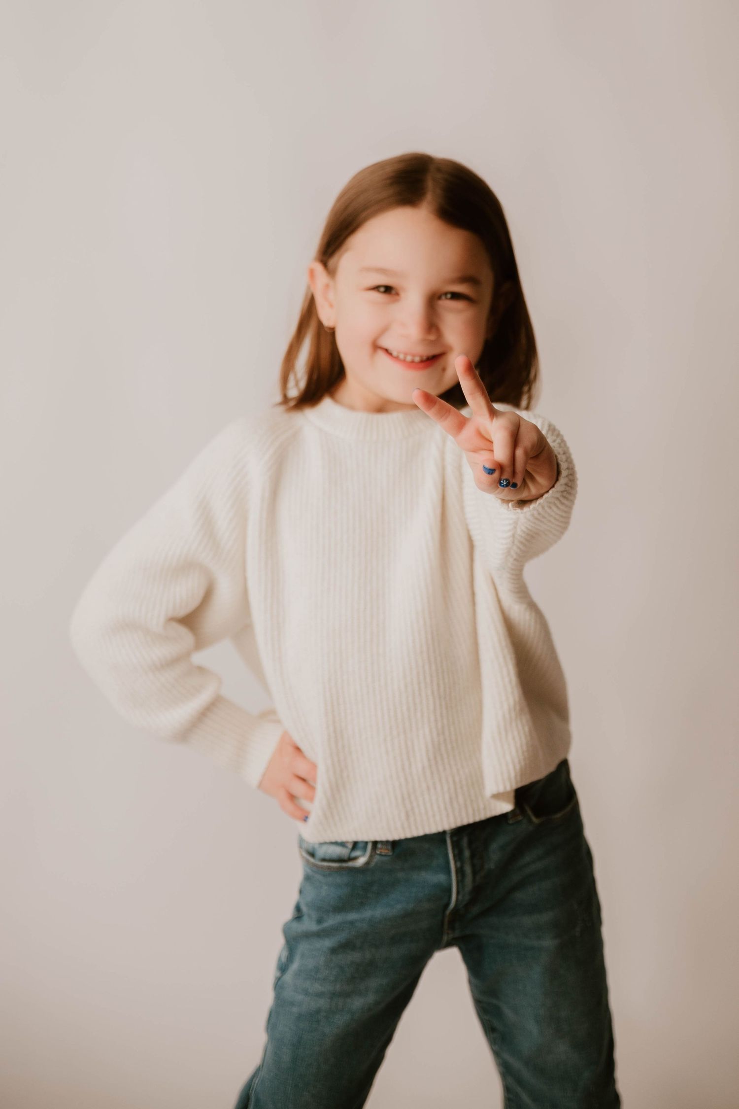 A young child in a white sweater and jeans poses playfully against a neutral background with a peace sign gesture.