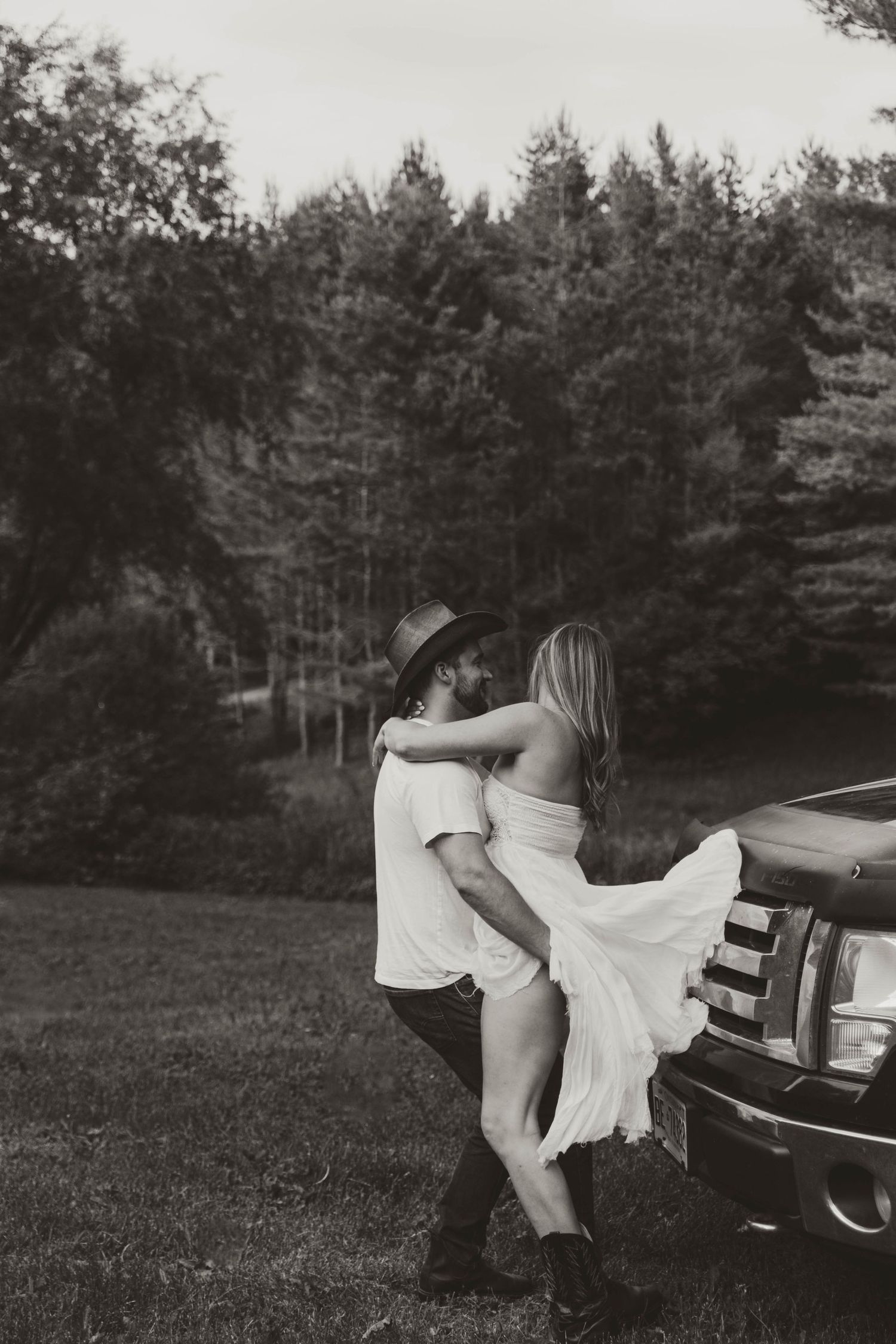 A romantic black and white photo of a couple embracing by a vintage truck with pine trees in the background.