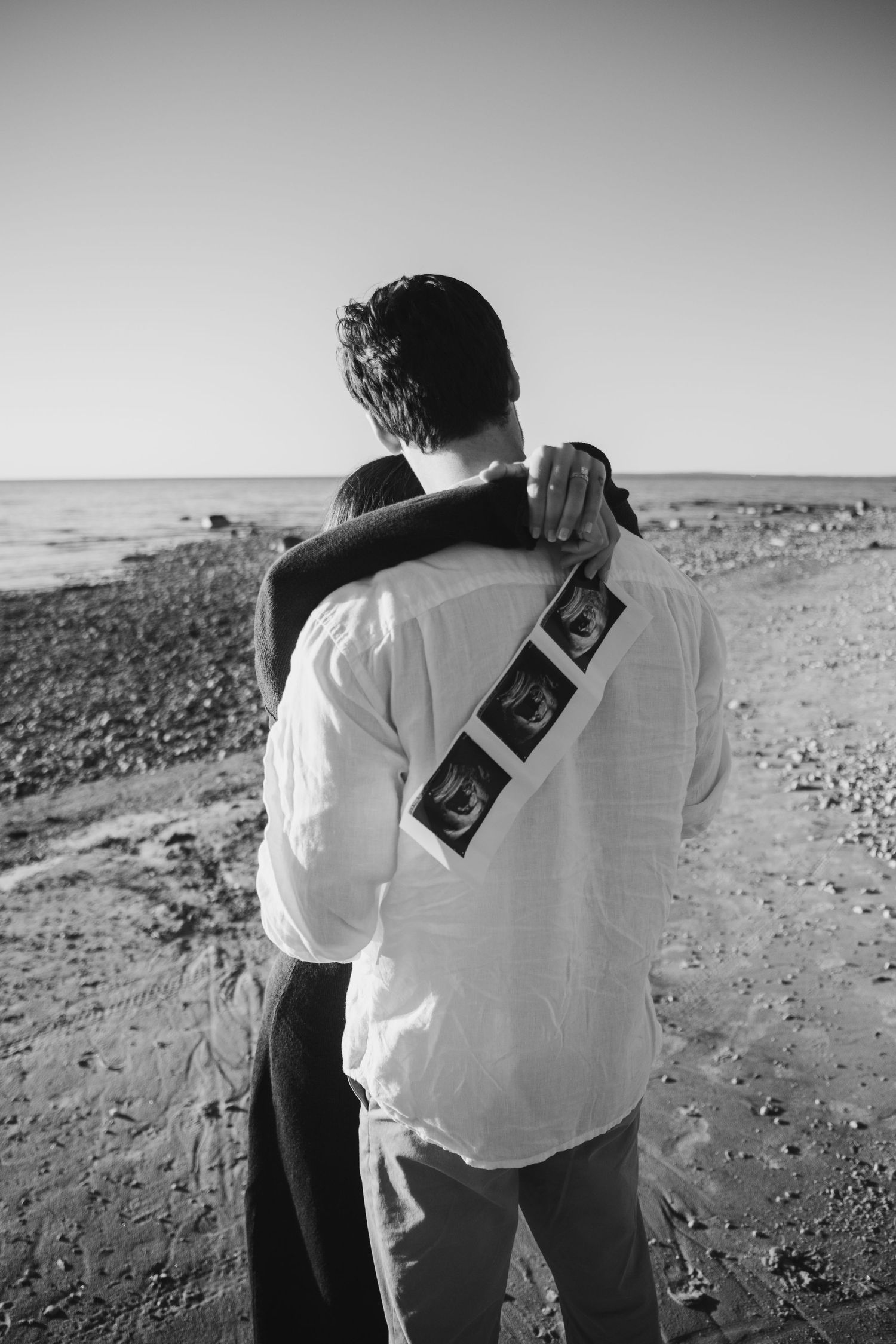 Black and white photo of a couple embracing on a pebbly beach, with ultrasound pictures draped over the man's shoulder.