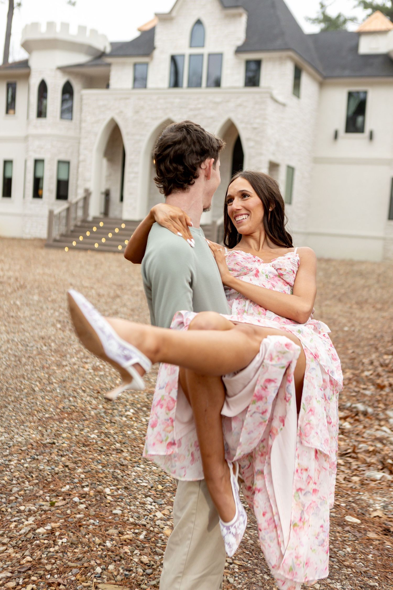Man lifting woman in floral dress outside elegant white mansion, romantic couple photo.