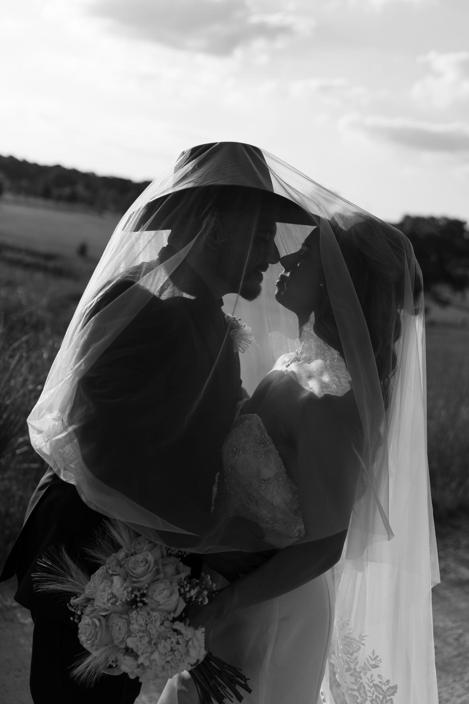 Black and white photo of bride and groom sharing intimate moment under wedding veil outdoors.
