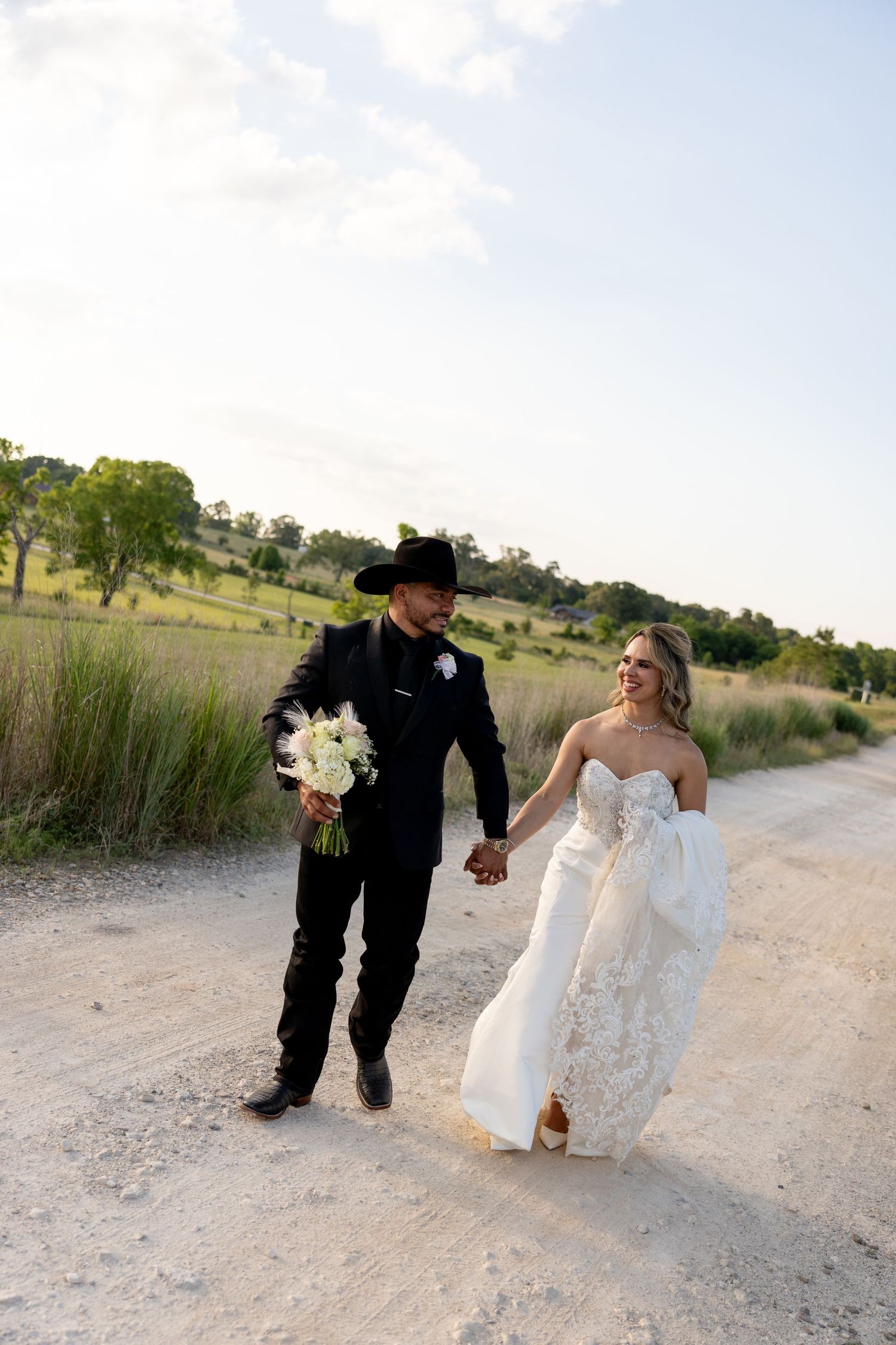 Bride and groom walk hand-in-hand on a dirt path, groom in black cowboy hat holding white bouquet.
