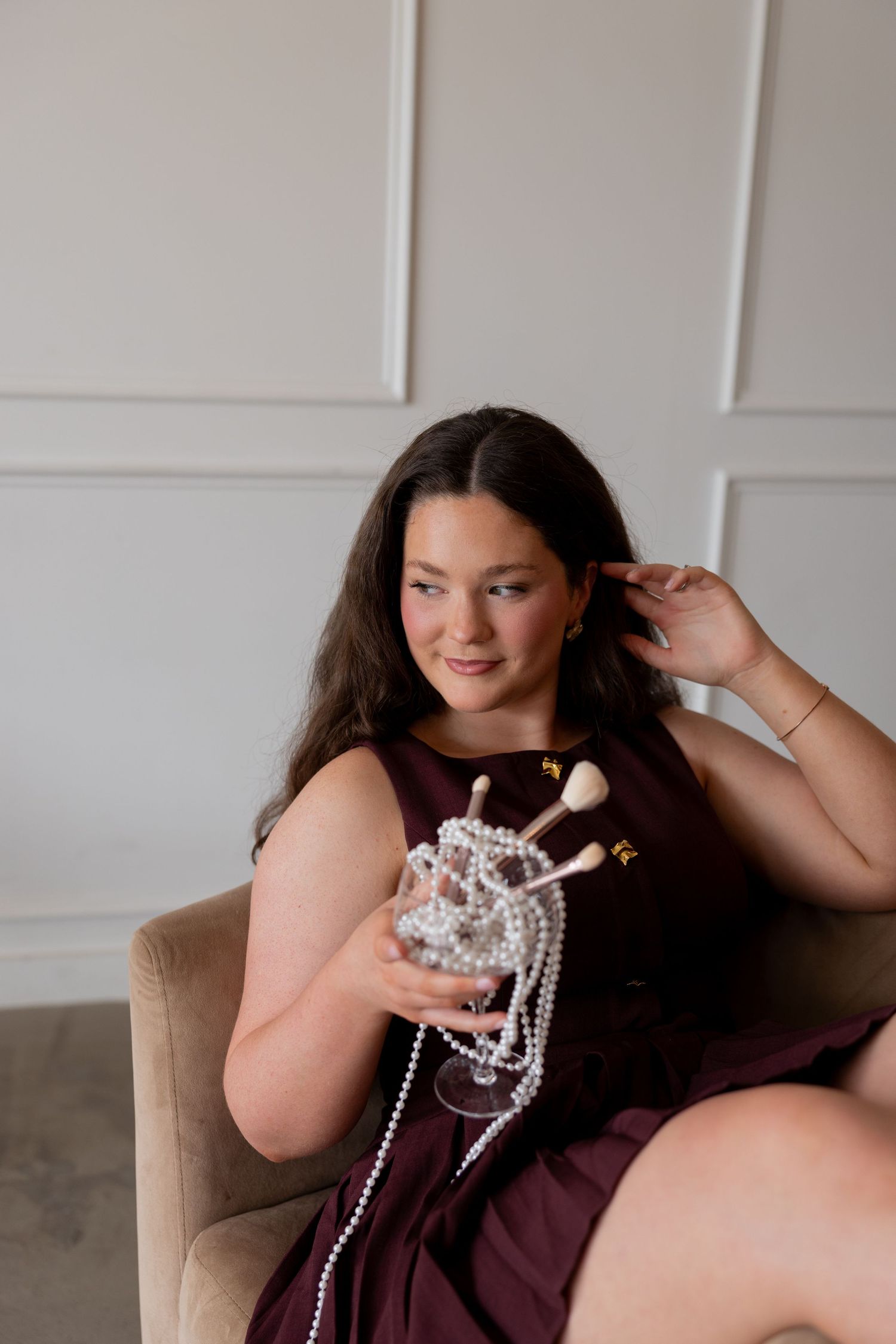 Woman in burgundy dress sitting in chair holding makeup brushes and pearl necklace in a wine glass.