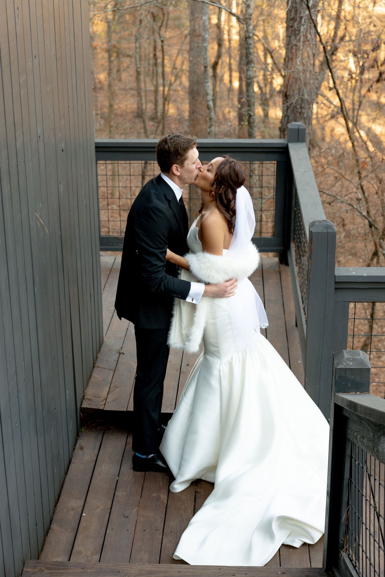Bride and groom share a romantic kiss on a wooden deck surrounded by autumn trees, bride wearing white gown and fur wrap.