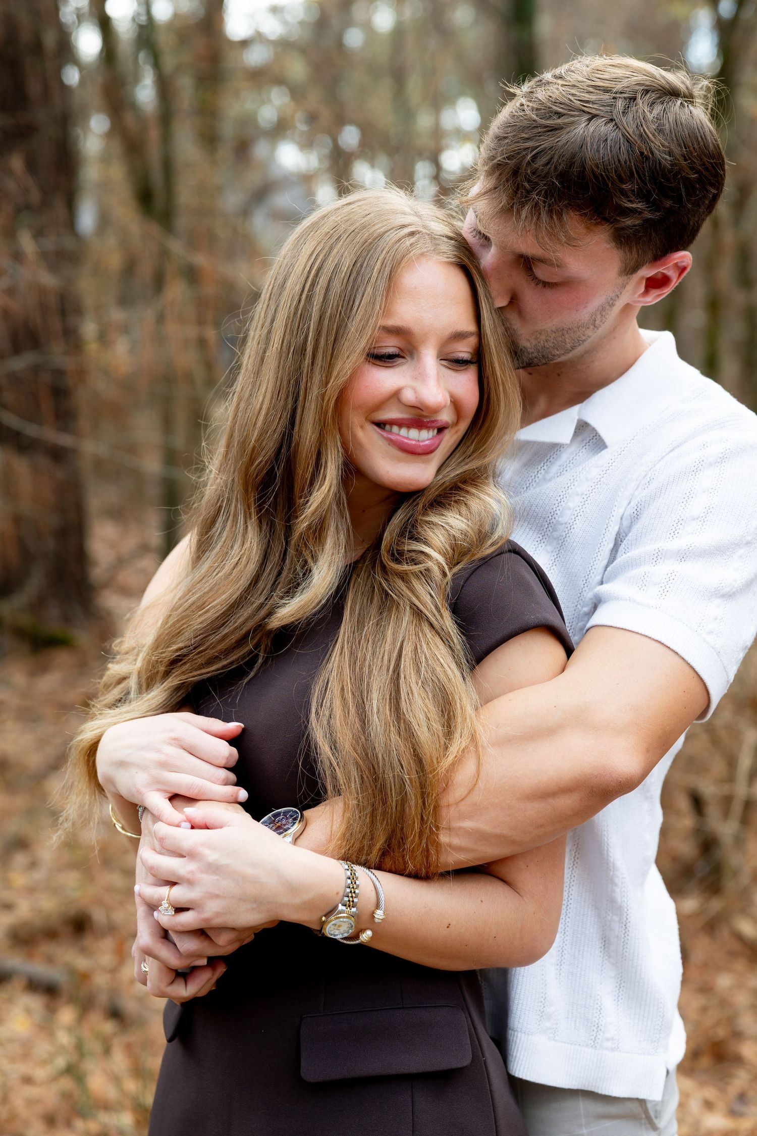 A romantic couple embraces and smiles together during an outdoor photo session in a wooded setting.