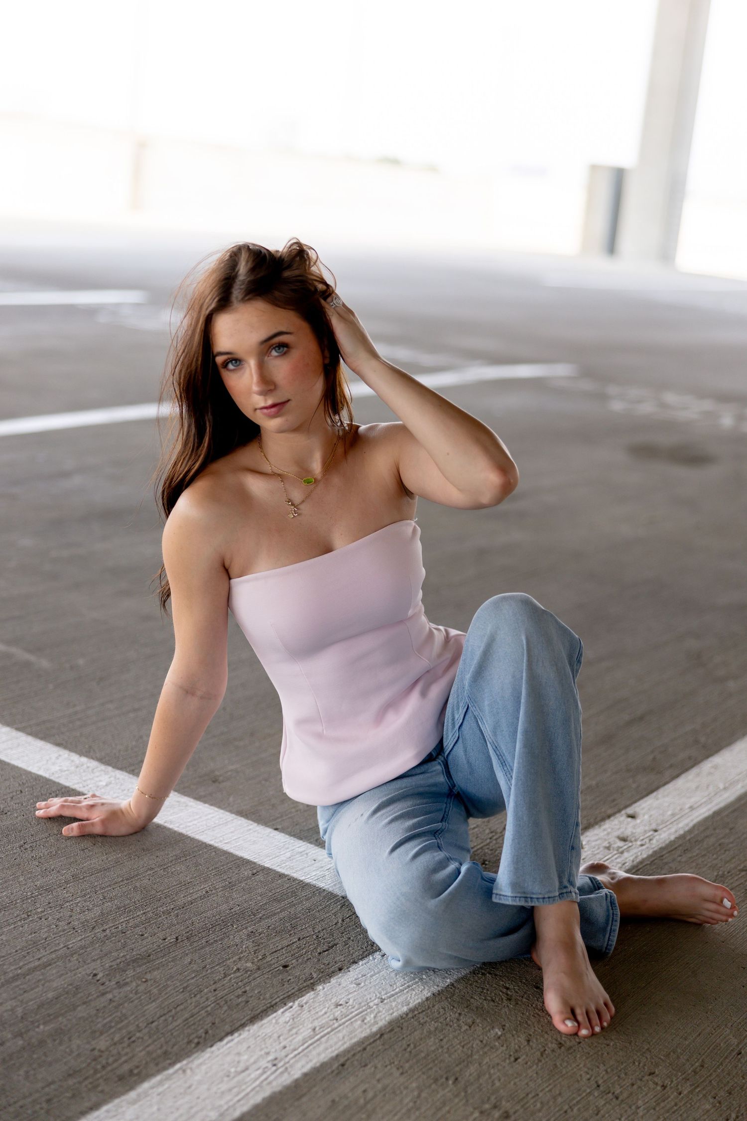 Young woman in pink tube top and jeans sitting barefoot on parking garage floor, posing with hand in hair.