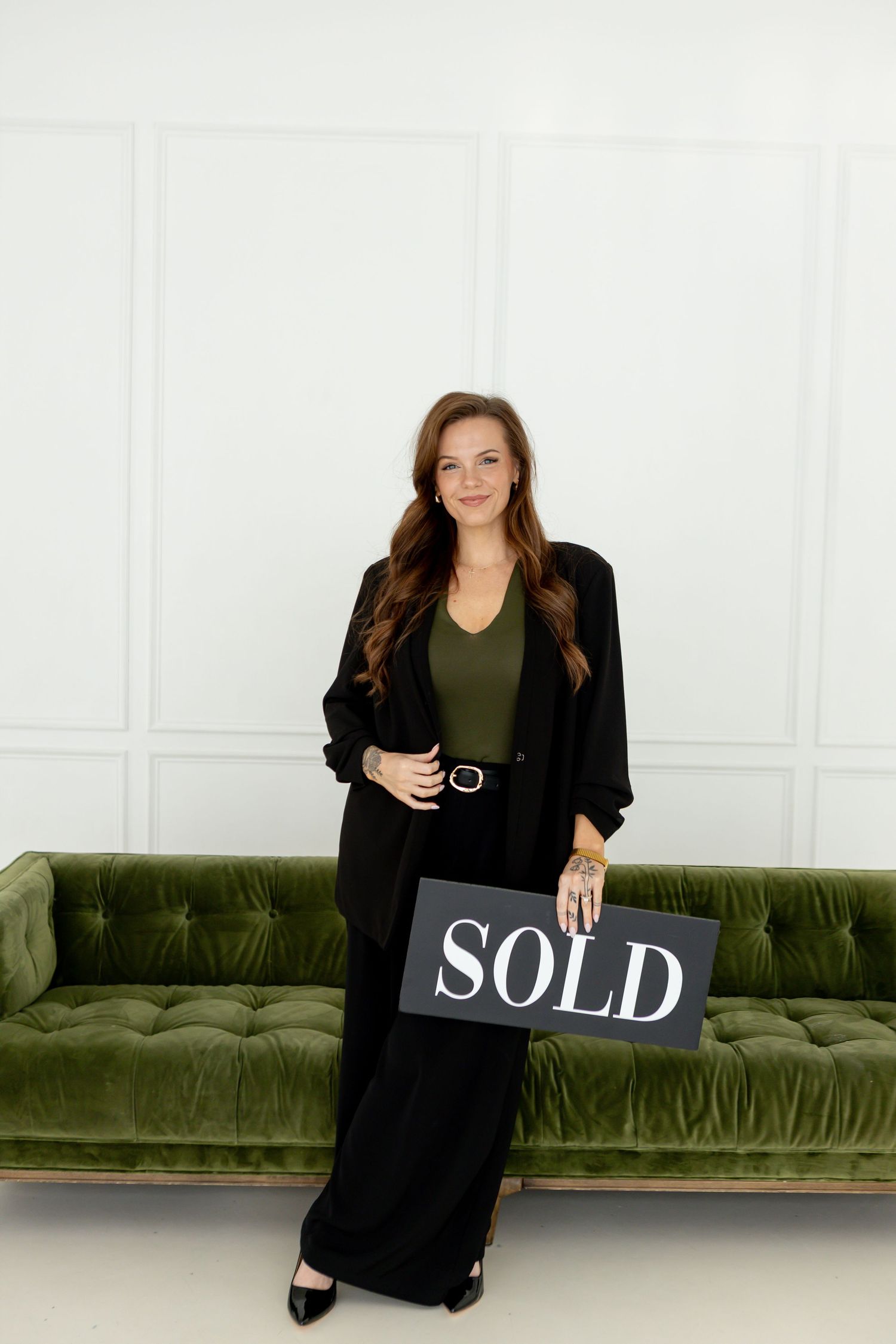 A smiling woman in black outfit holds a SOLD sign, standing before a green velvet sofa in a bright white room.