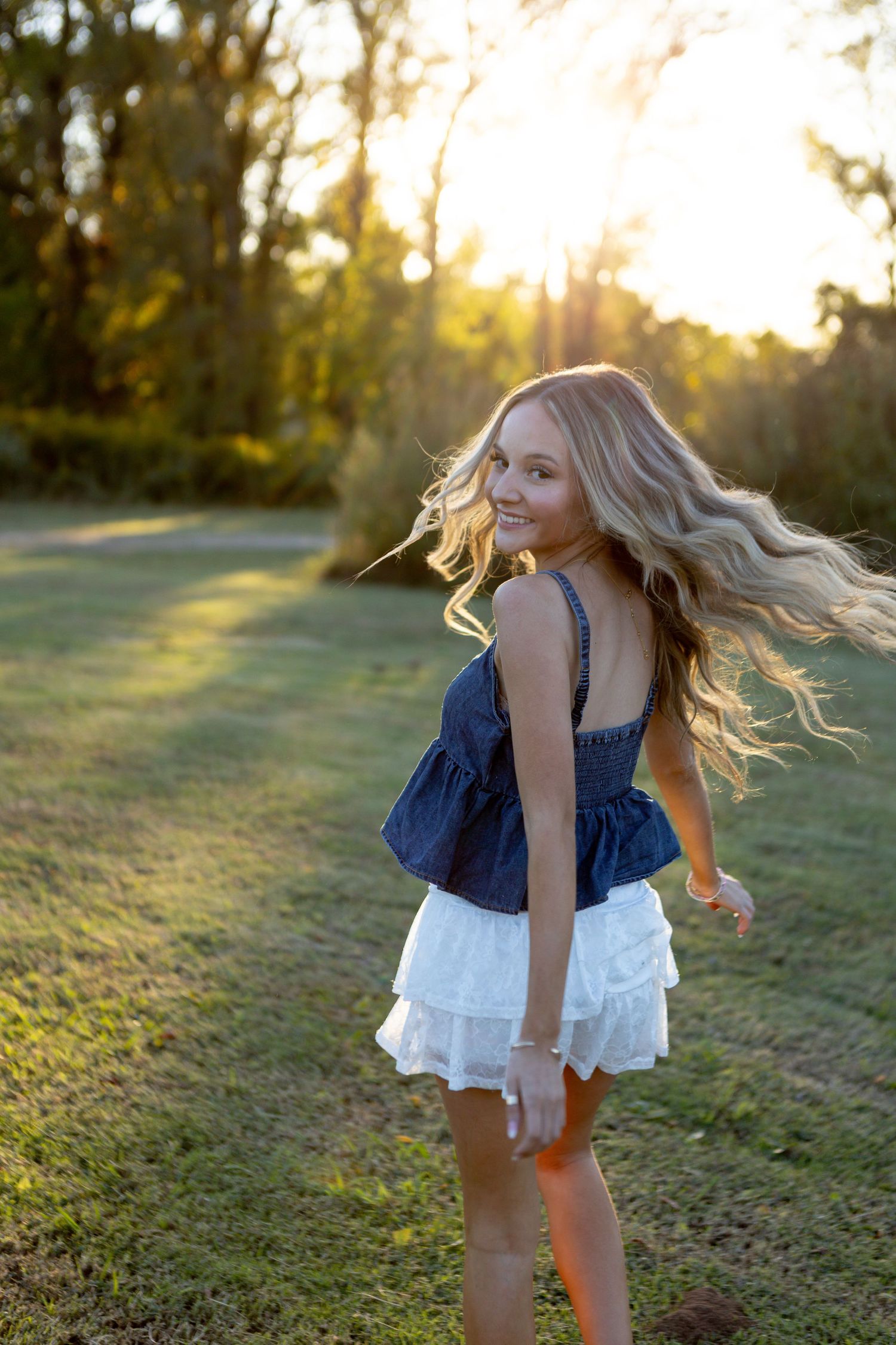 Blonde girl in denim top and white skirt twirling outdoors at golden hour sunset in a lush green park.