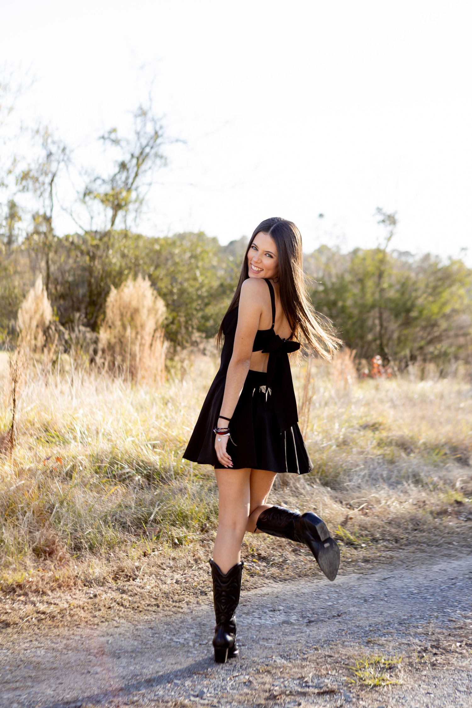 Young woman in black mini dress and cowboy boots smiling on a rural dirt path surrounded by dry grass.