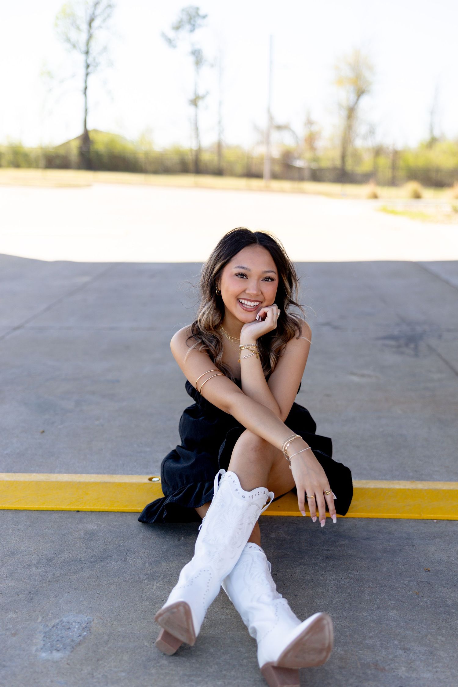 Young woman in black dress and white cowboy boots sitting on a curb, smiling outdoors on a sunny day.