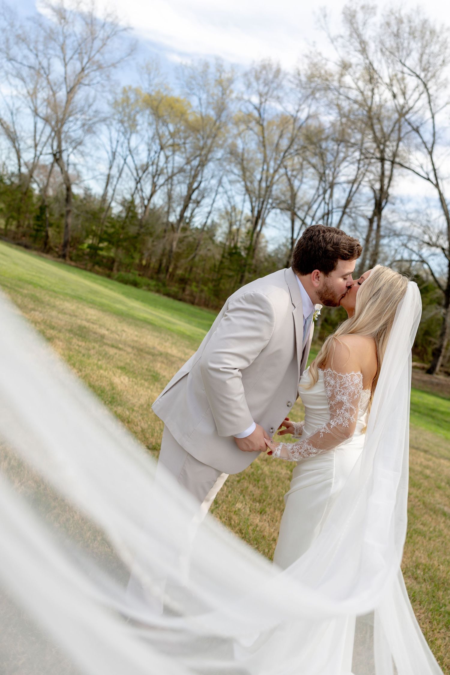 Bride and groom share a romantic kiss outdoors, with her flowing veil billowing in the breeze against a lush green backdrop.