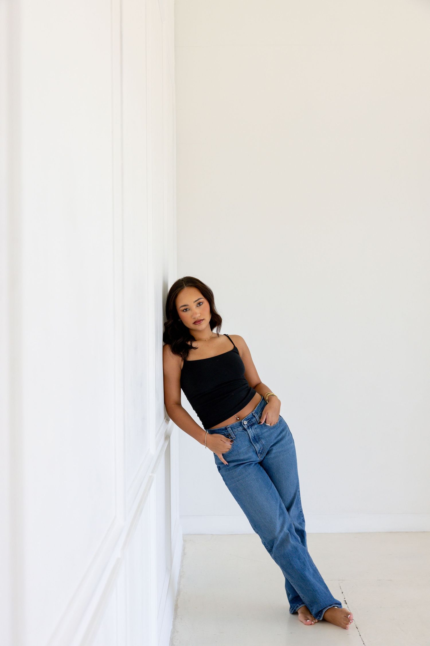 Young woman in black crop top and blue jeans leaning against white wall, casual fashion portrait.
