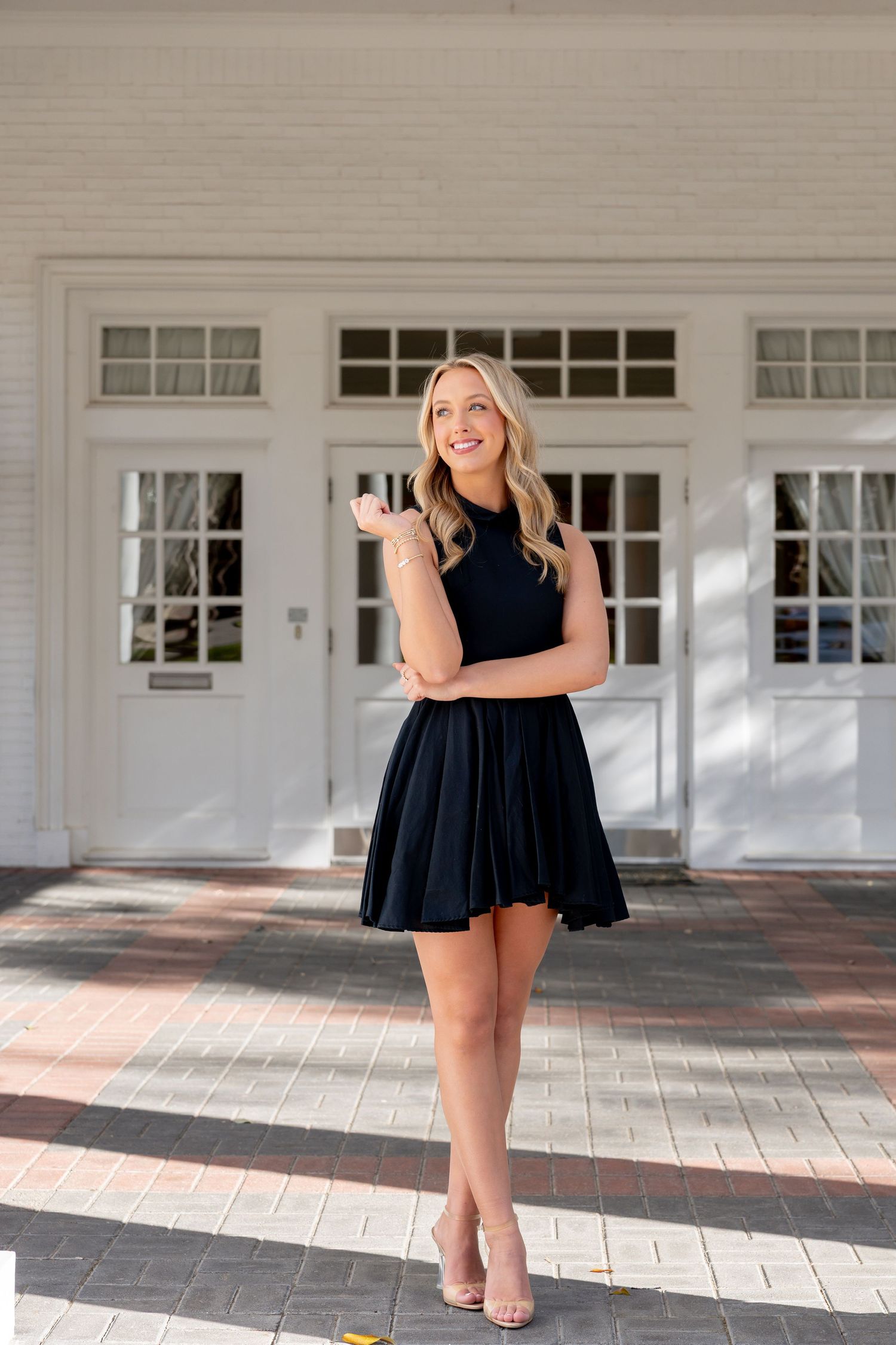 Young woman in black mini dress and nude heels posing confidently in front of white building doors.