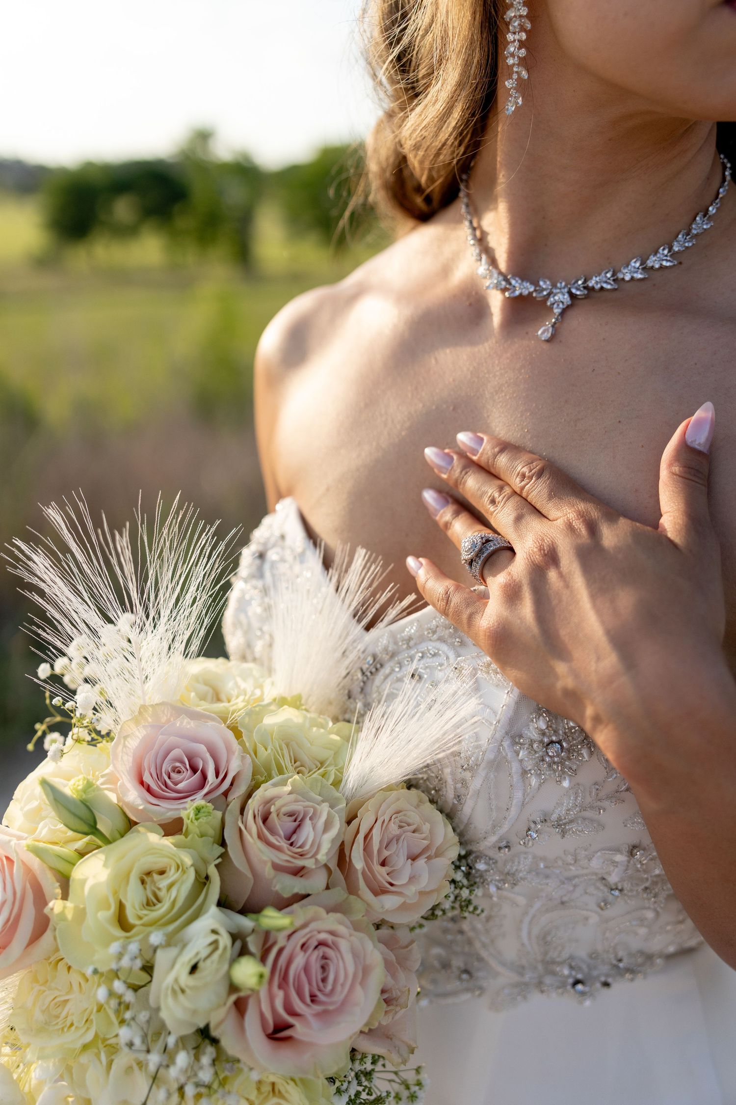Bride in white gown holding pink rose bouquet with feathers, wearing diamond necklace and ring, hand on chest.