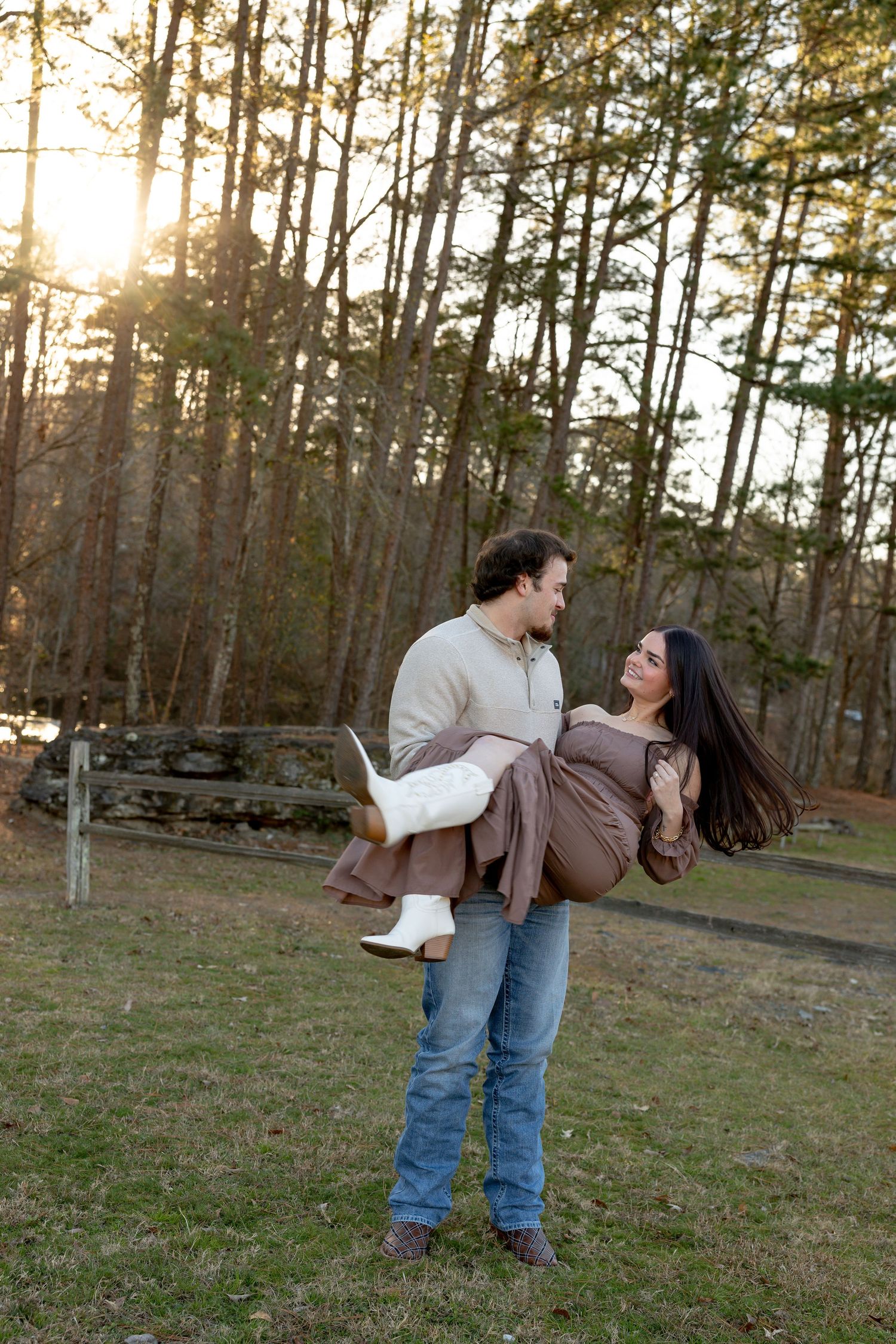 A man carries a smiling woman in his arms outdoors at sunset, surrounded by tall trees in a romantic setting.