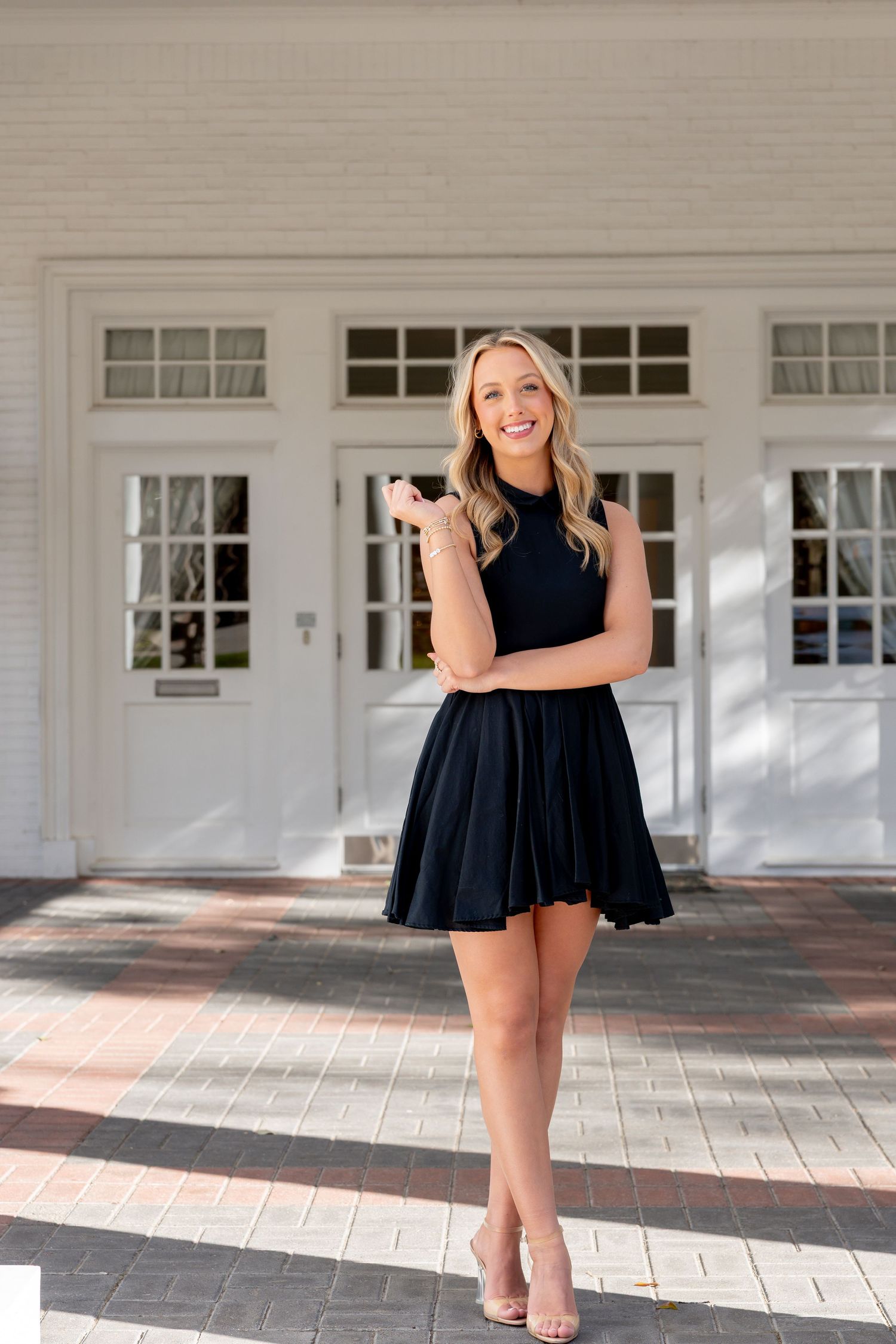 Smiling blonde woman in a navy blue fit-and-flare mini dress and nude heels posing outdoors near white building.