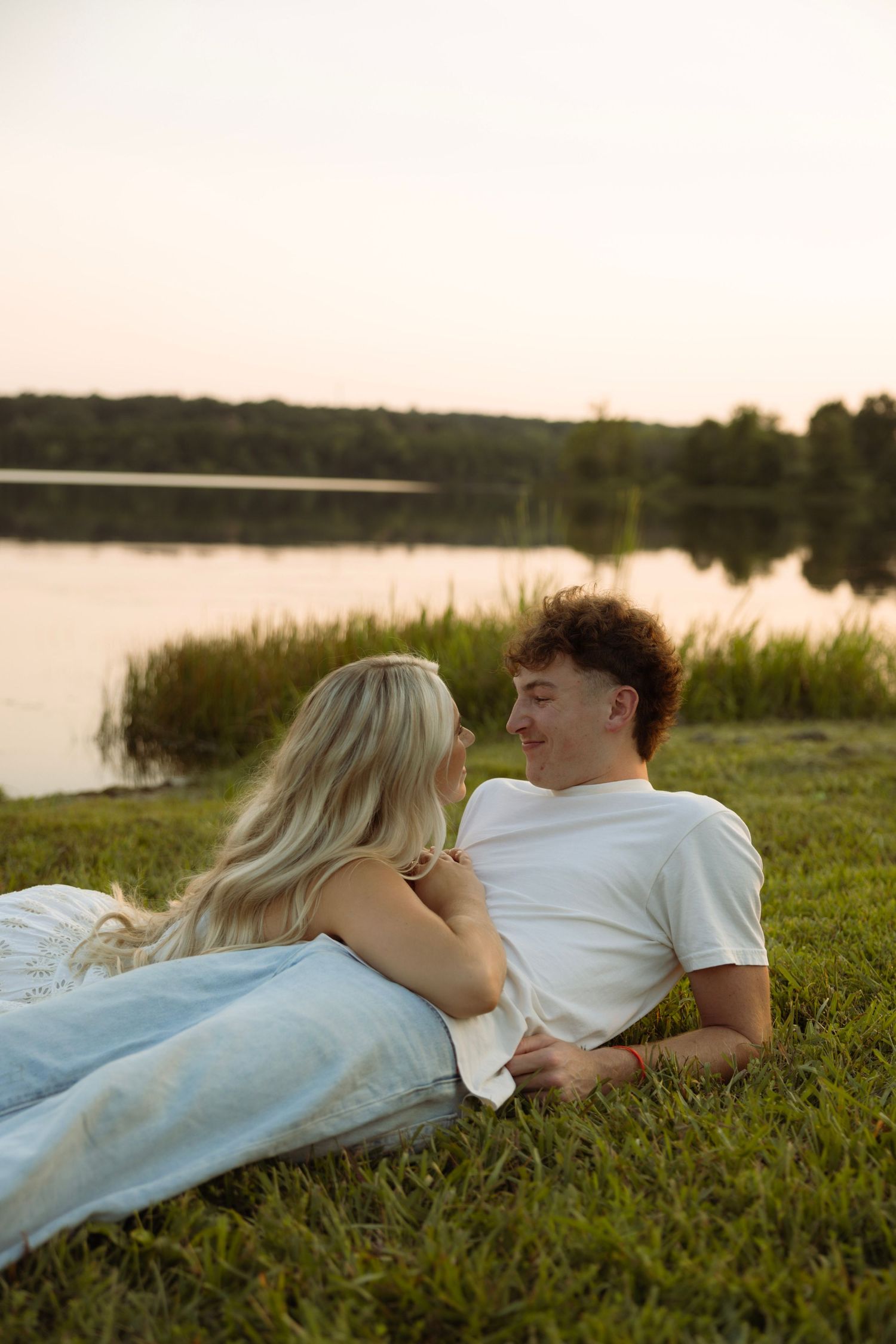 Couple lying on grass by a lake at sunset, smiling and gazing at each other romantically.