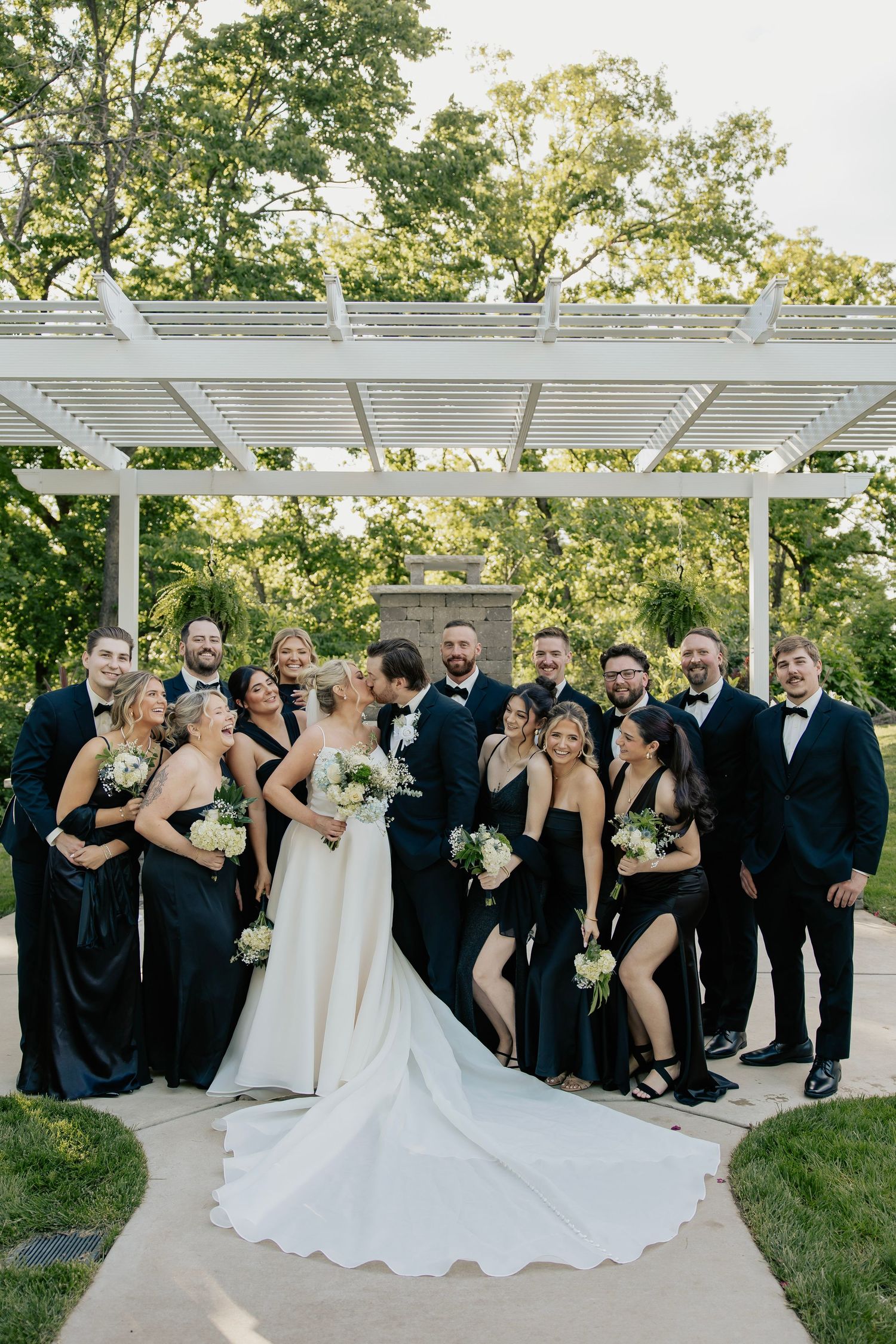 Wedding party in black attire poses with bride and groom under white pergola surrounded by lush greenery.
