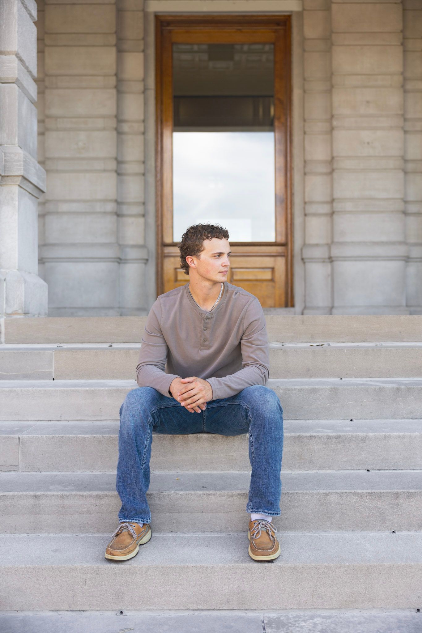Young man in a brown henley and jeans sitting on stone steps in front of a classic building.
