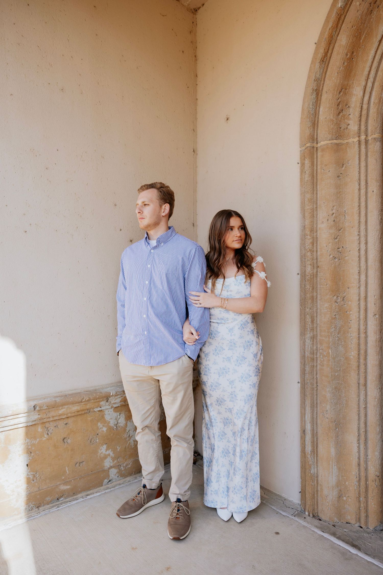 Engaged couple standing together against a warm beige stone wall in soft natural light.