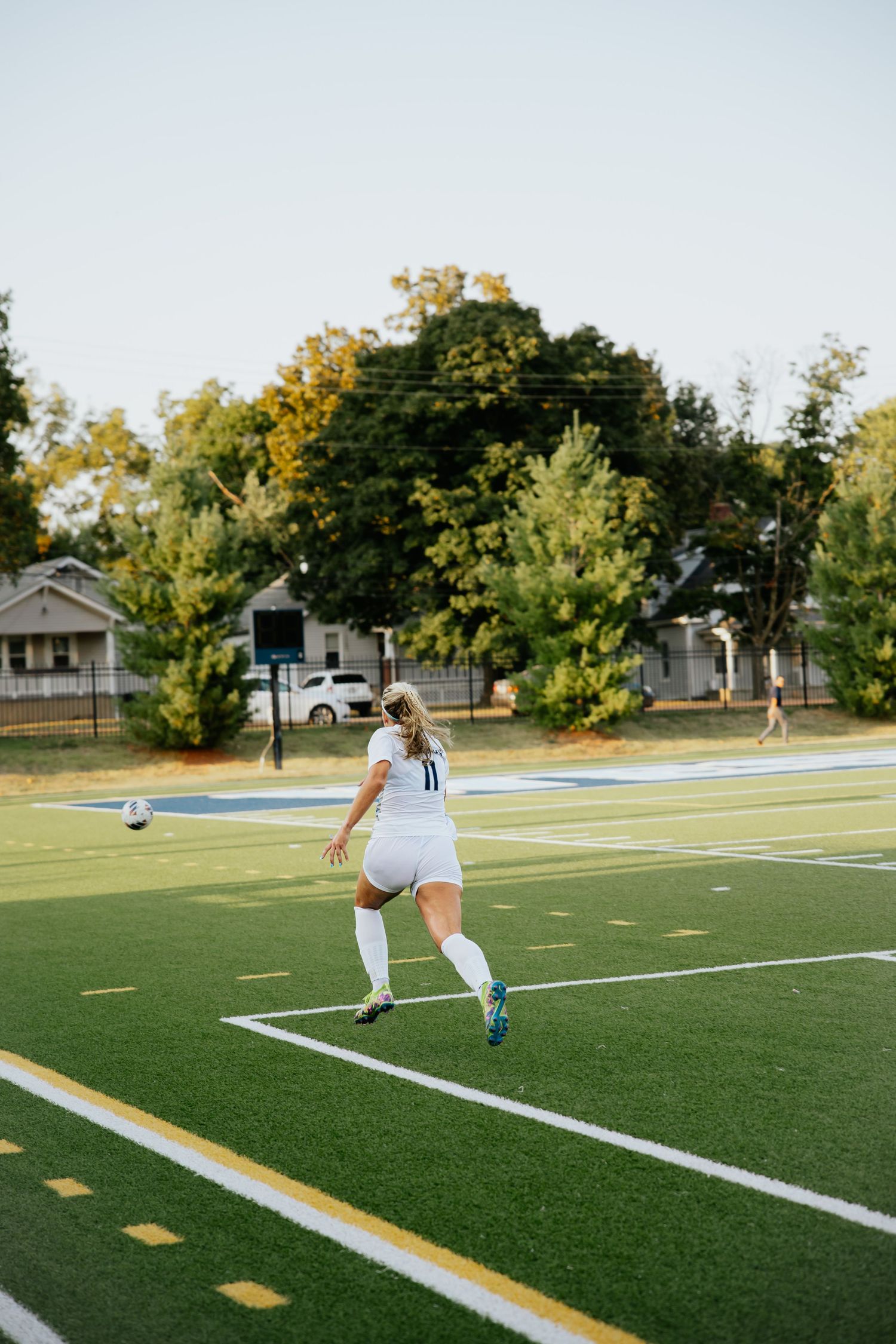 Female soccer player in white jersey #11 kicks ball on green turf field during sunset game.