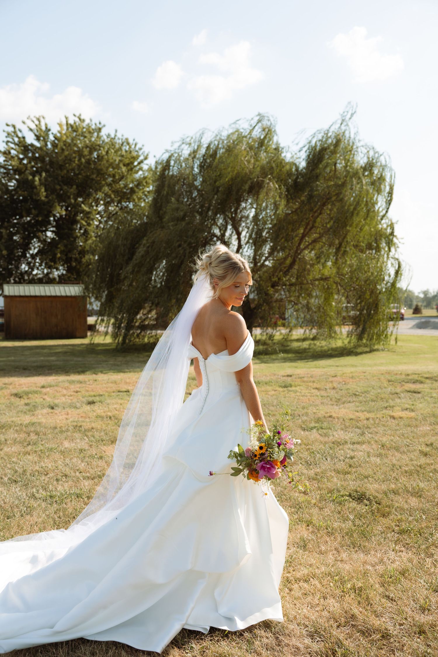 Bride in white off-shoulder gown with veil and colorful bouquet stands outdoors near weeping willow tree.