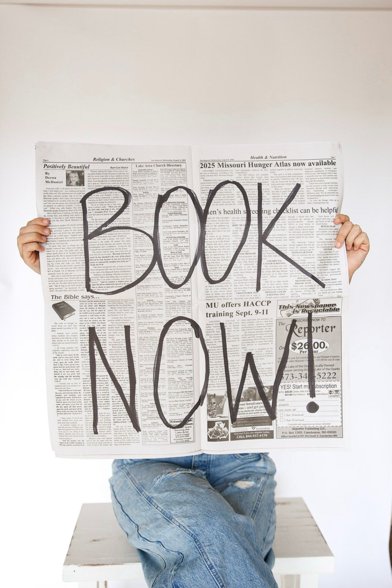 Person holding newspaper with bold handwritten text reading BOOK NOW exclamation point.