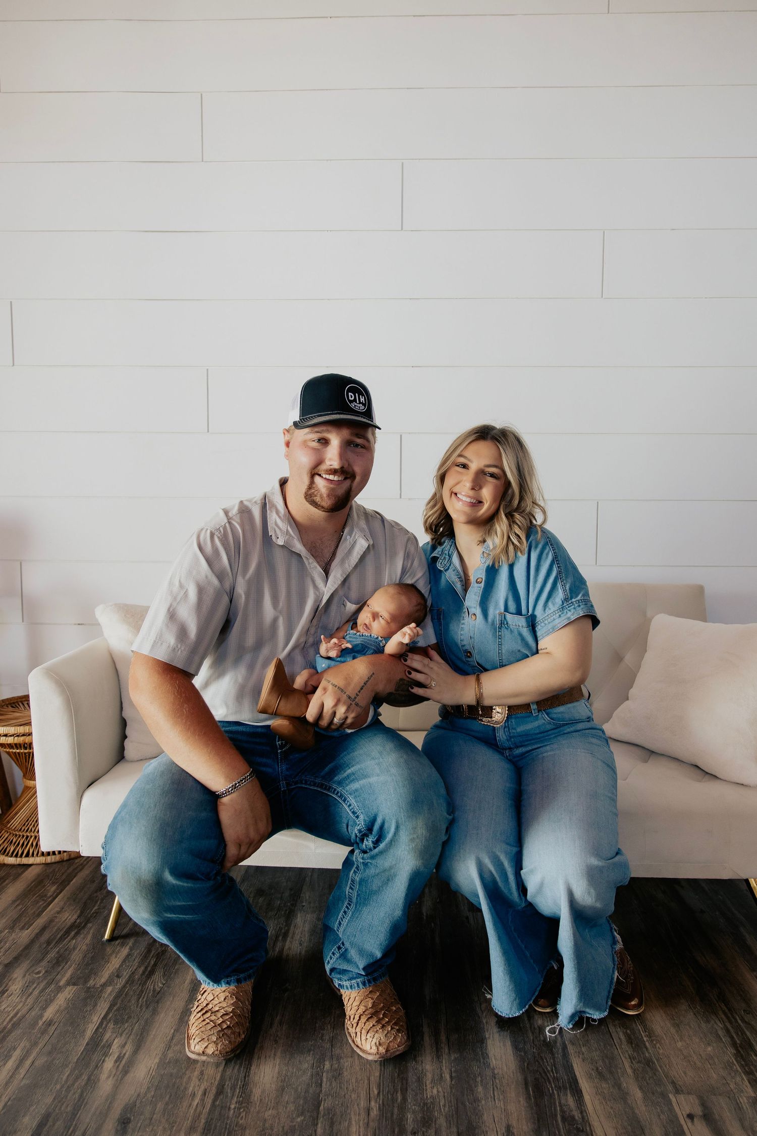 A smiling couple sits on a white couch holding their newborn baby, dressed in casual denim outfits in a bright studio.