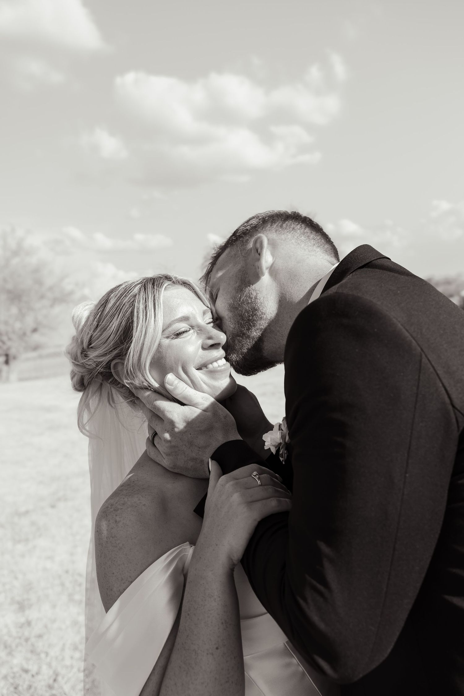 Black and white photo of a groom kissing bride on cheek as she smiles joyfully outdoors on their wedding day.