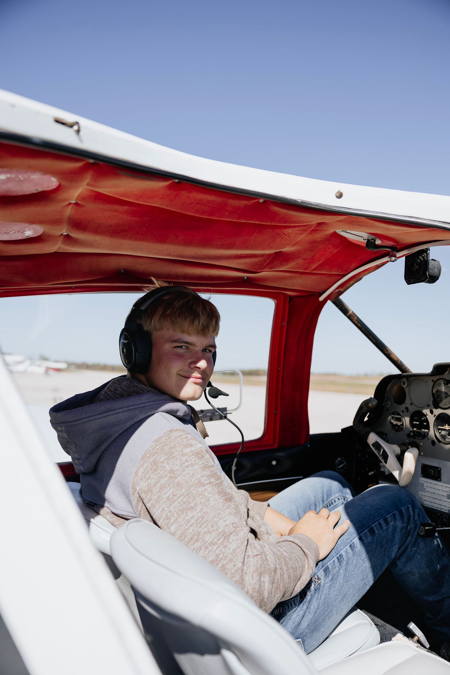 Teen boy wearing headset sitting in cockpit of small red airplane.