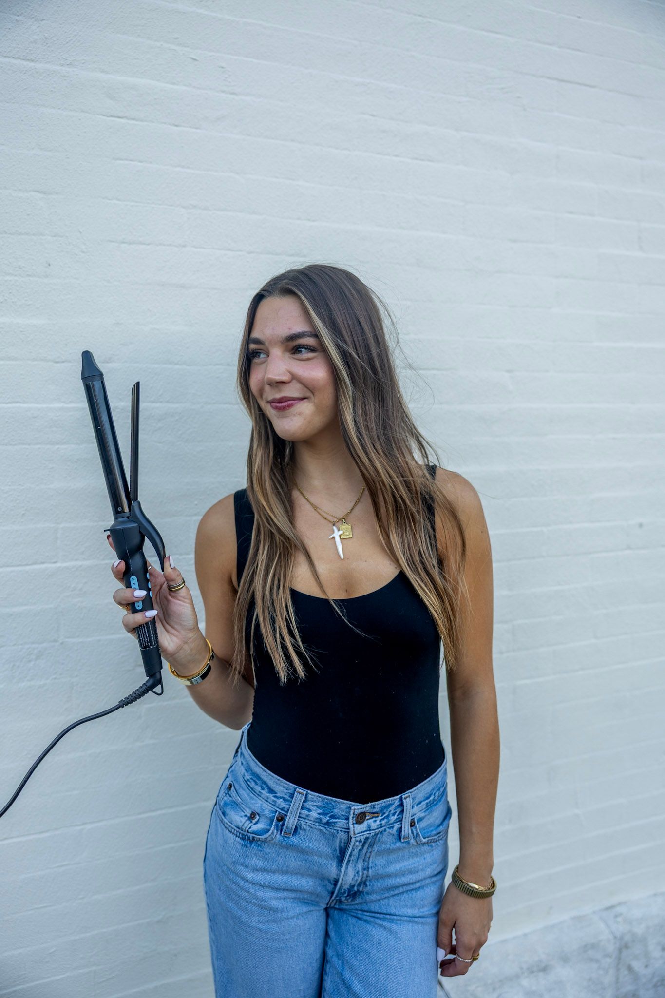 A young woman in a black bodysuit holds a curling iron while posing against a white brick wall outdoors.