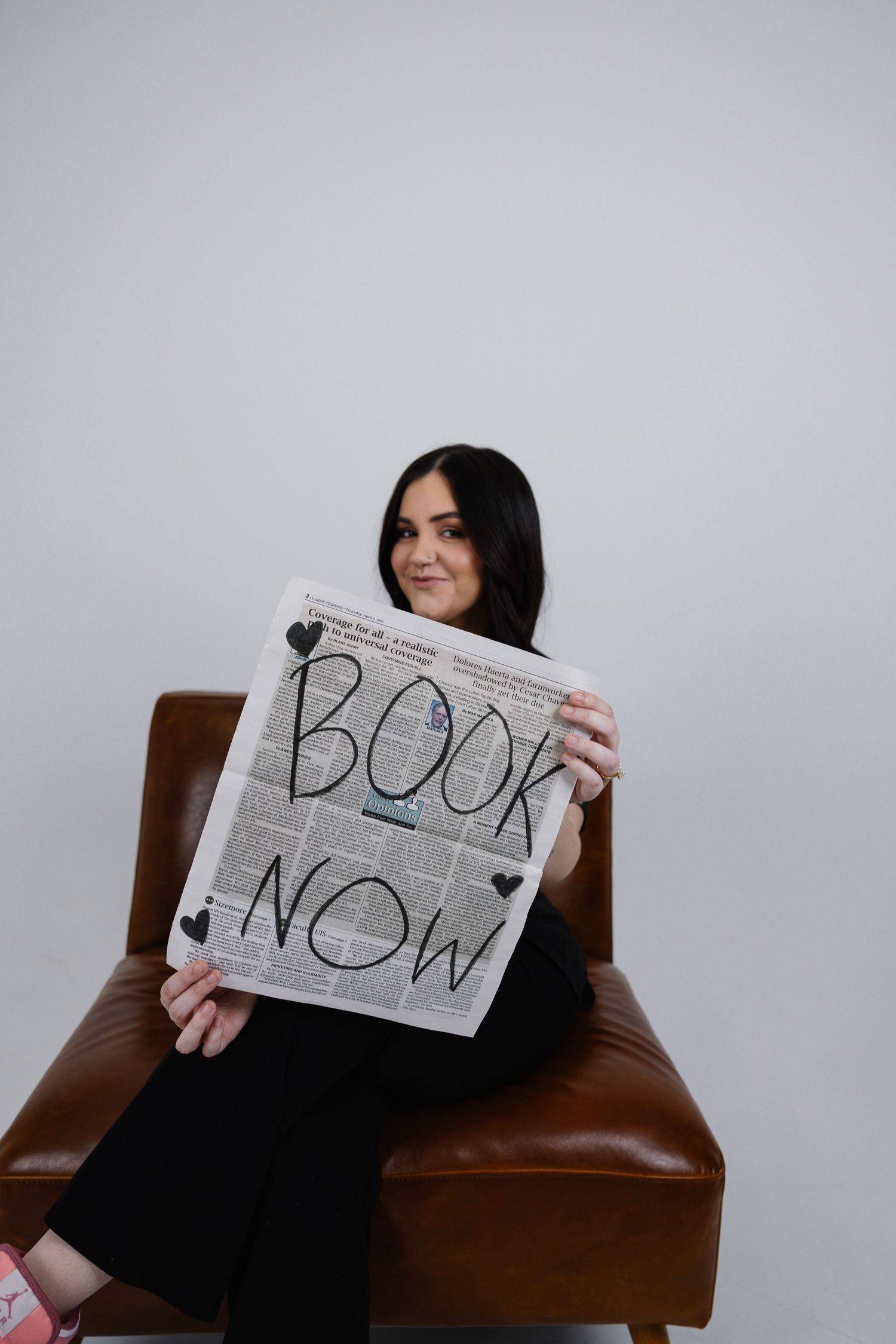 Dark-haired woman sitting in leather chair holding newspaper with BOOK NOW written on it.