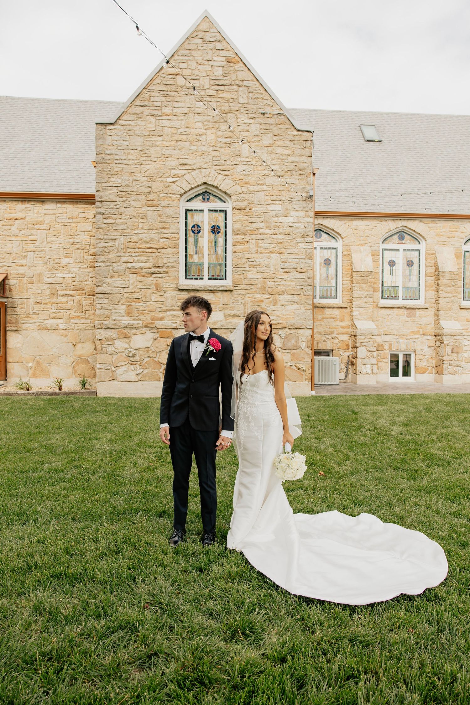 Bride and groom holding hands standing on green grass in front of a stone church on their wedding day.