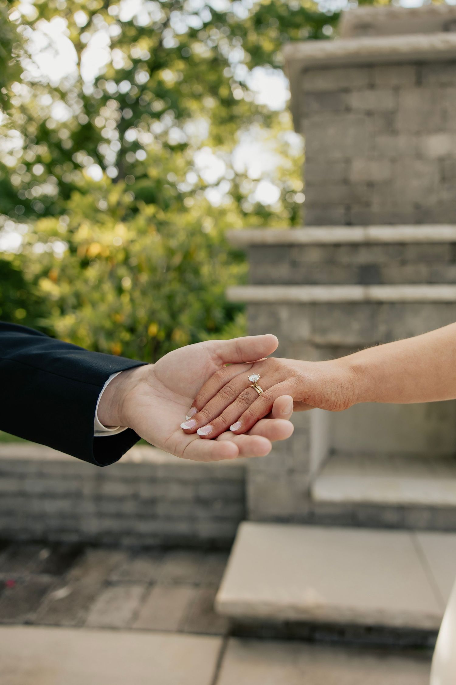 Newlywed couple holding hands outdoors, showcasing wedding rings, with stone steps and green trees in the background.