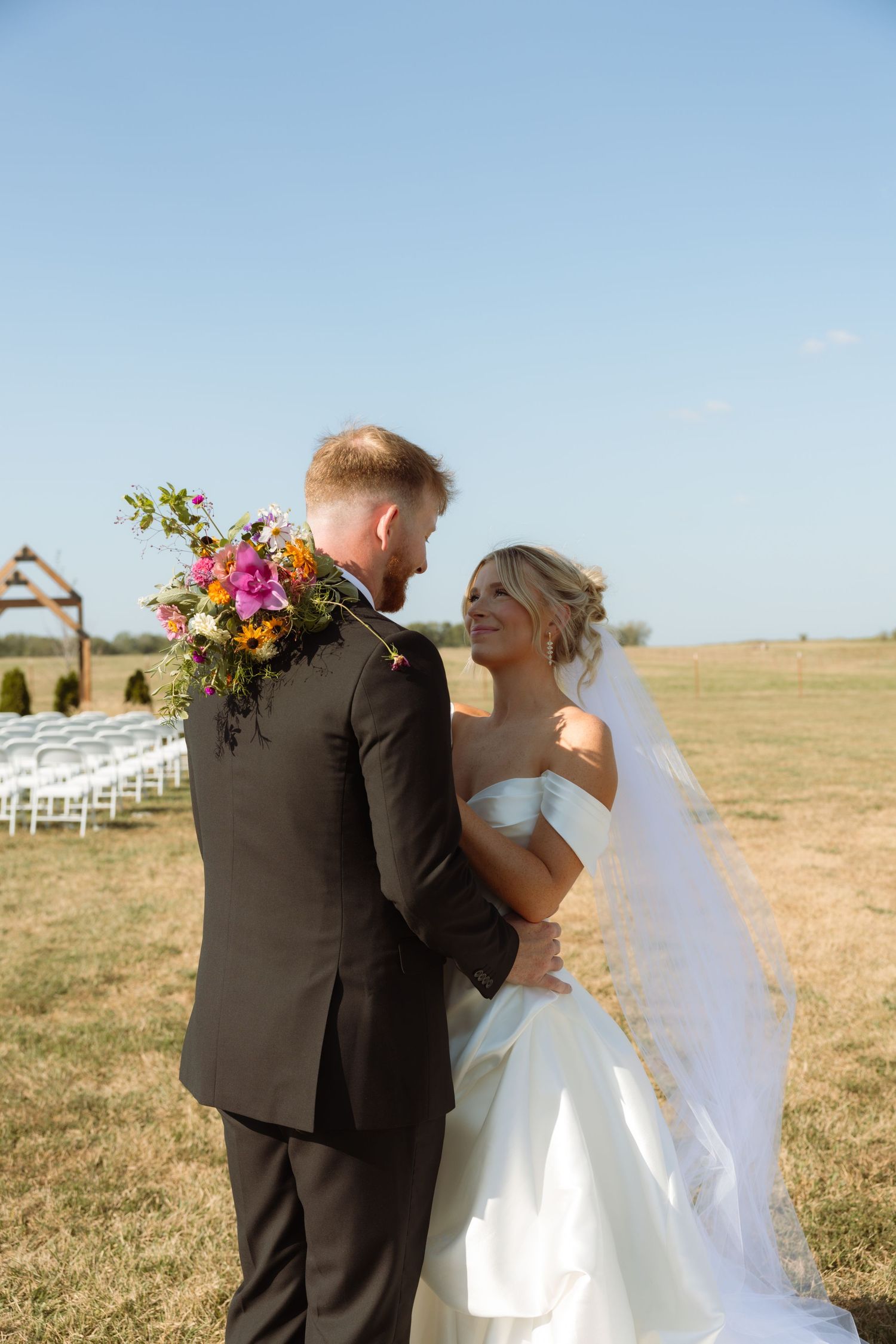 A smiling bride in a white gown holds colorful flowers while embracing her groom in a dark suit at an outdoor wedding.