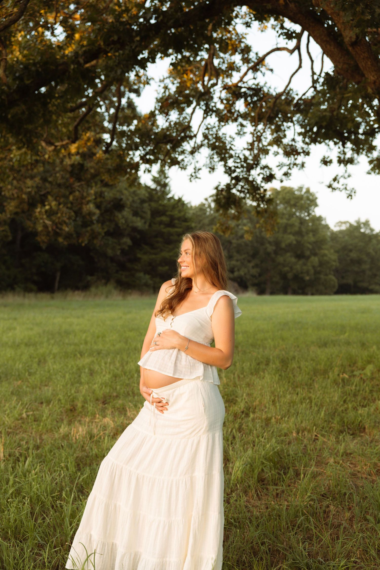 Pregnant woman in white dress standing in green field under oak tree at golden hour sunset.