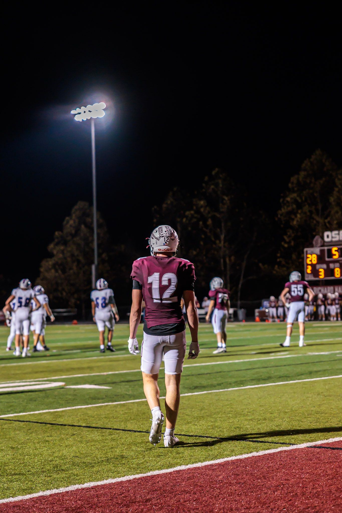 Football player #12 in maroon uniform walks on field during night game under bright stadium lights.