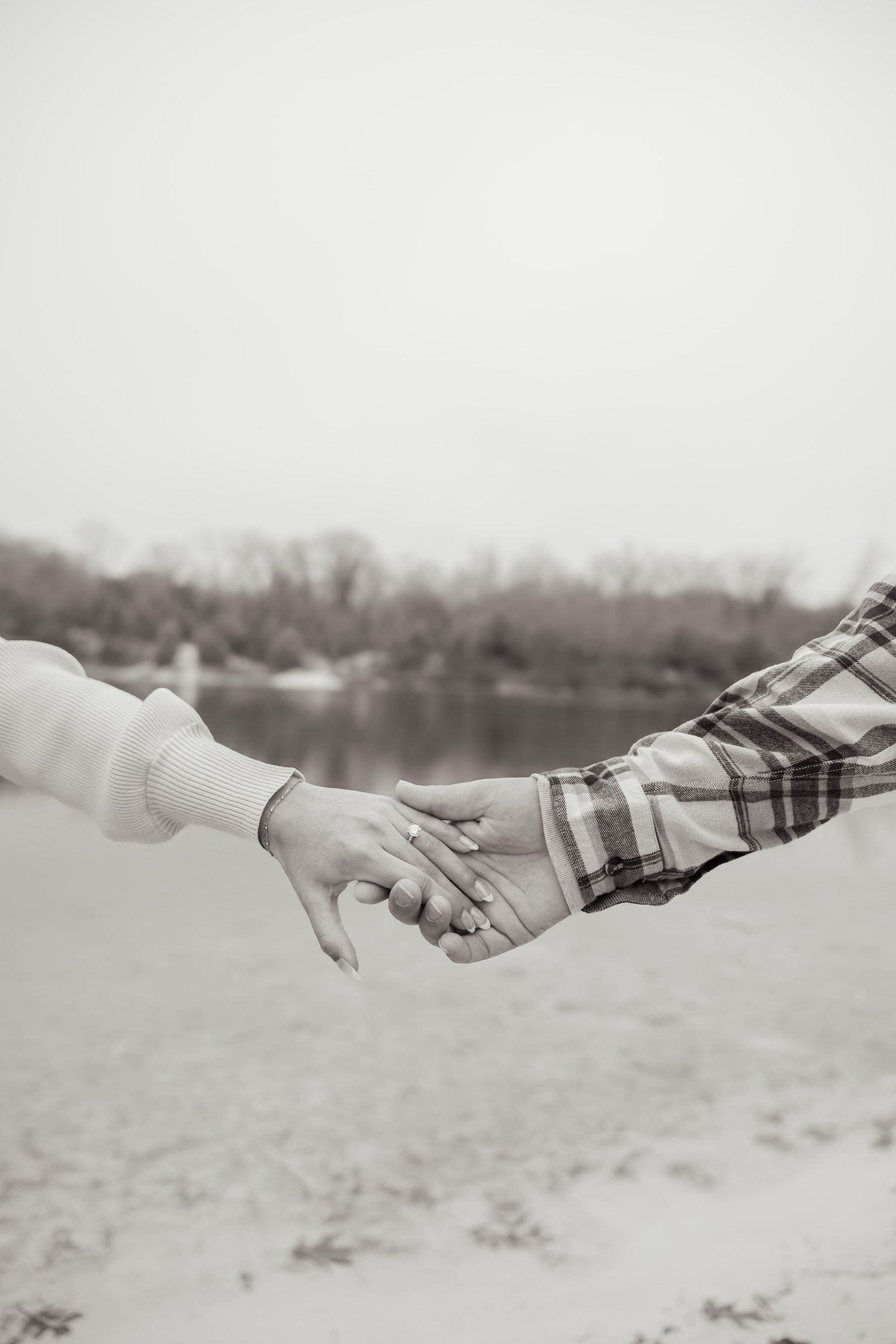 Black and white close-up of a couple holding hands near a serene lakeside beach.
