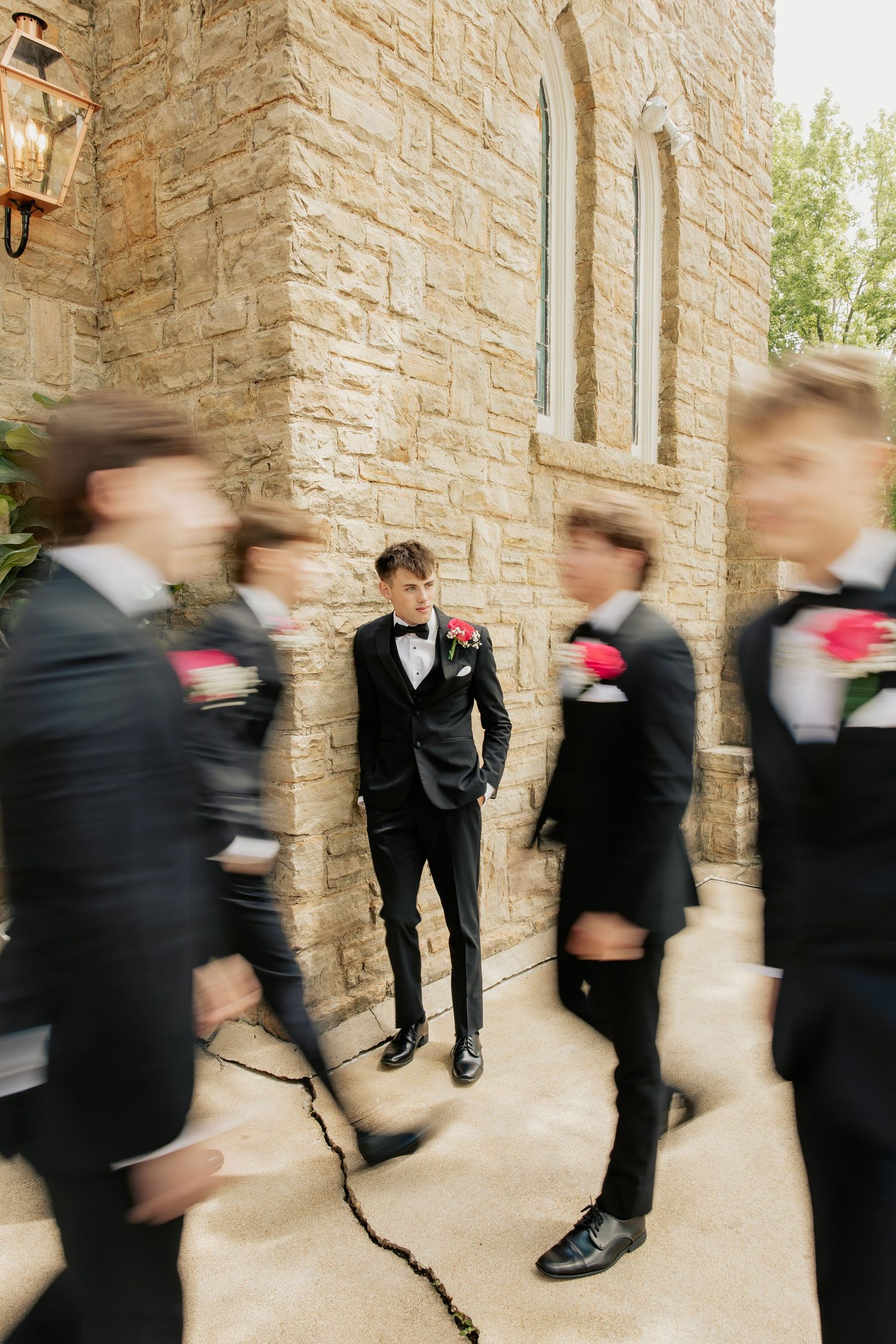 Creative motion blur photo of groomsmen in black tuxedos walking past groom outside stone venue.