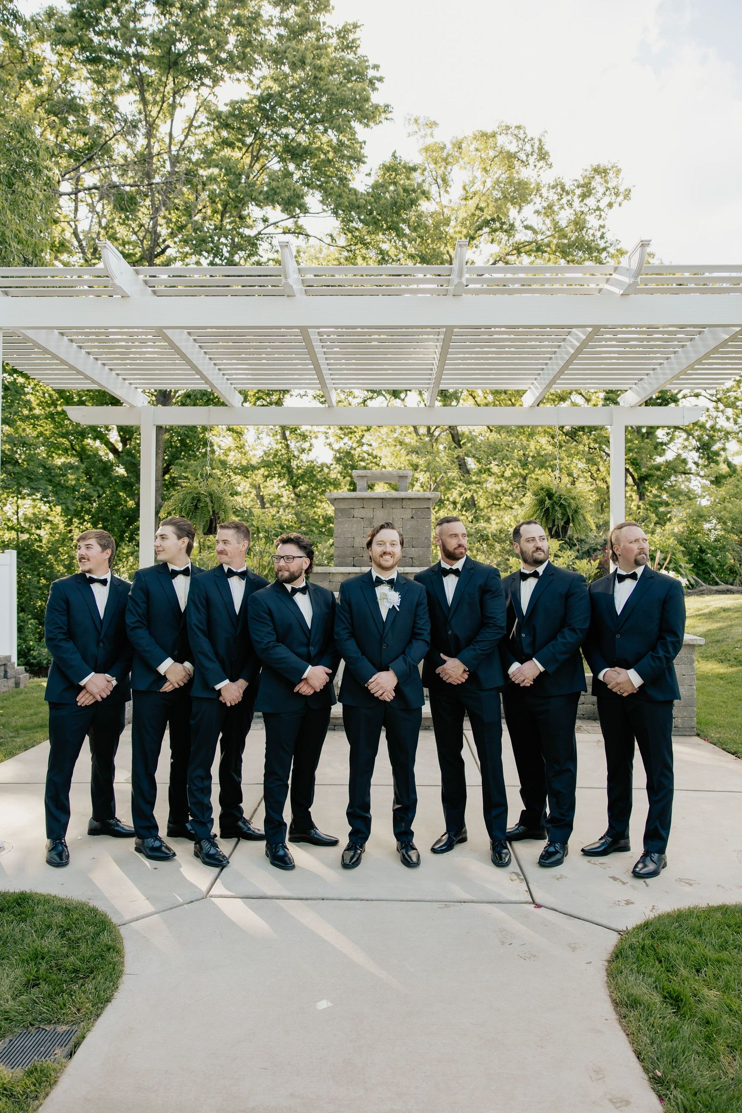 Groom and groomsmen in black tuxedos with bow ties pose under a white pergola outdoors.