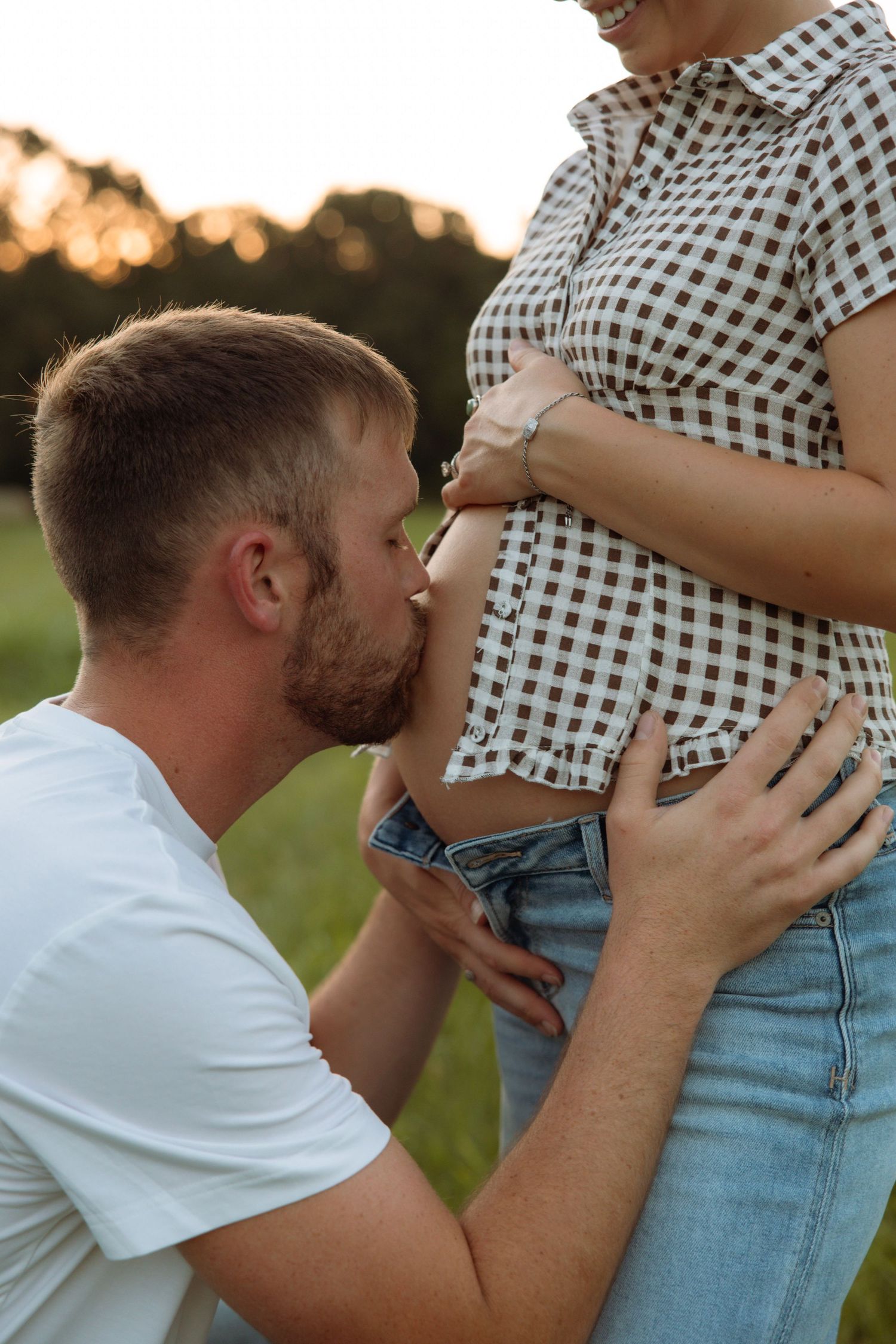 A man kneeling and kissing a pregnant woman's belly outdoors at sunset in a tender maternity moment.