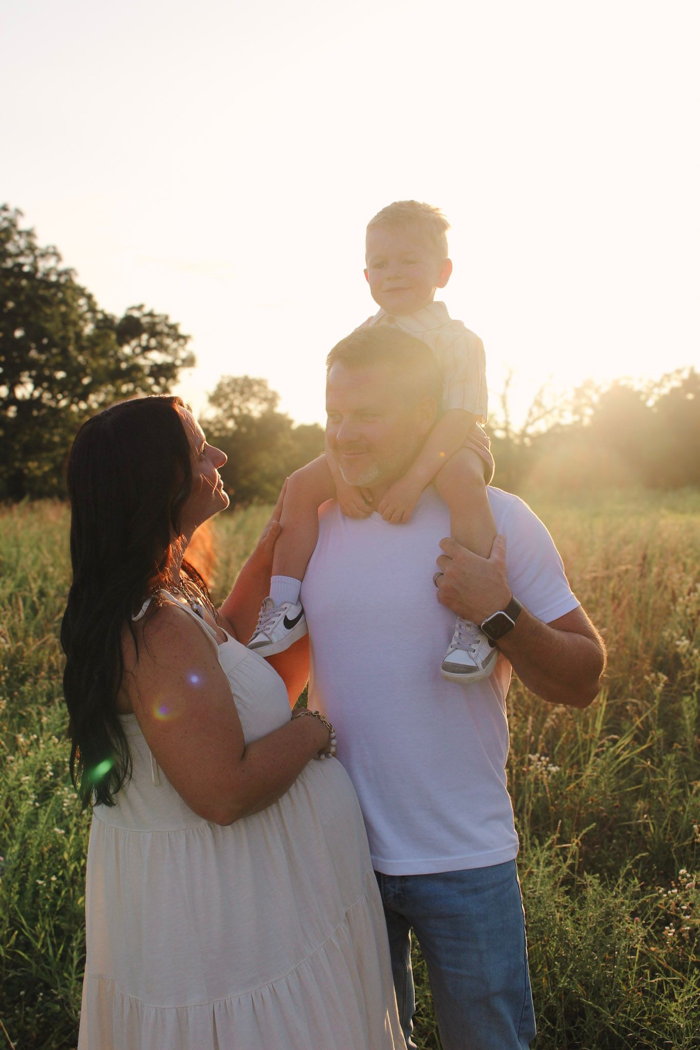 Happy family enjoys a sunny golden hour moment in a field, toddler perched on fathers shoulders.