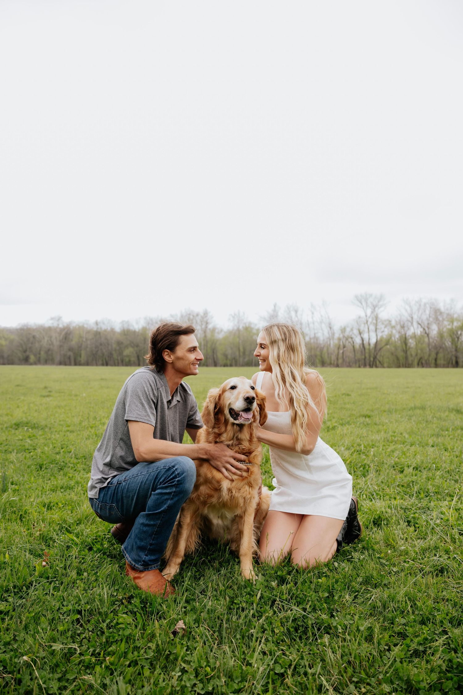Couple kneeling in green field with happy golden retriever dog on overcast spring day.